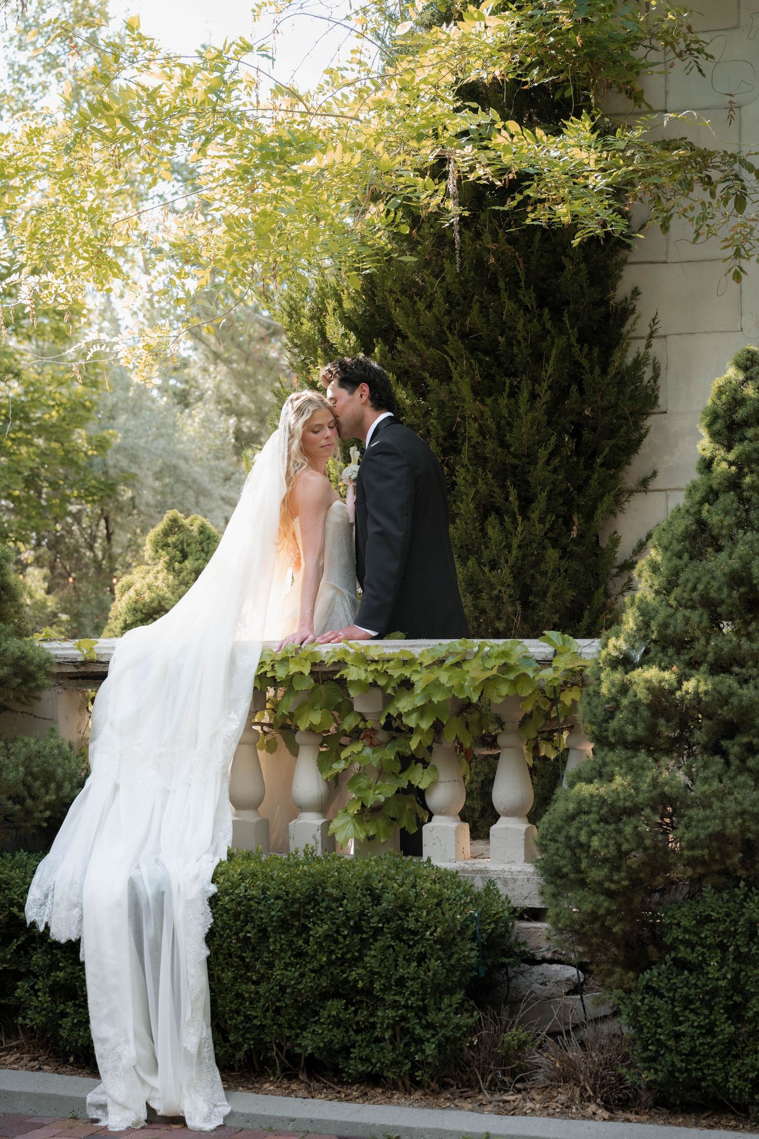 Bride and groom share a romantic kiss on an elegant stone balcony surrounded by lush greenery.