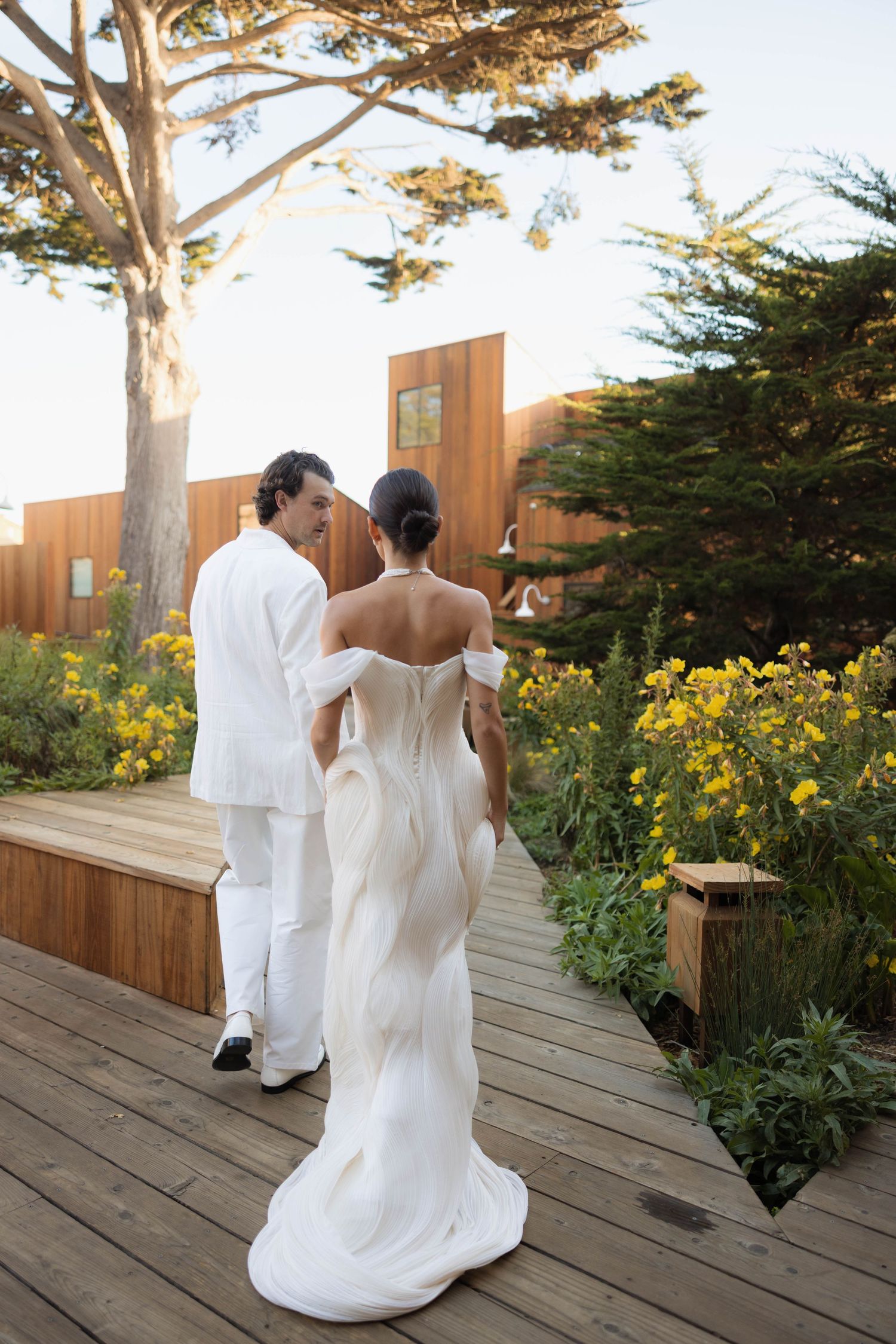 A bride in a white gown and groom in white suit walk hand in hand on a wooden deck surrounded by yellow flowers.
