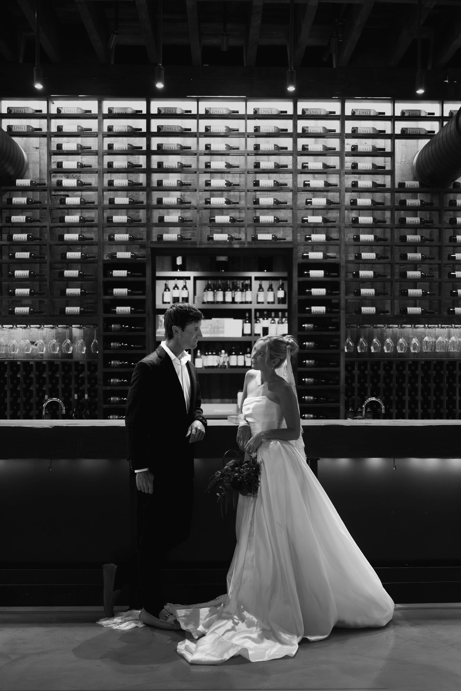 Black and white photo of a bride and groom standing before a large wine rack display on their wedding day.