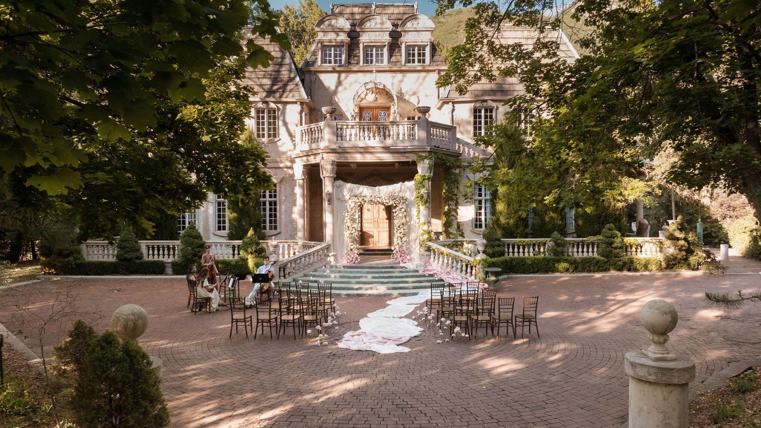 Charming stone manor house with outdoor wedding ceremony setup, white aisle runner, and surrounded by lush trees.