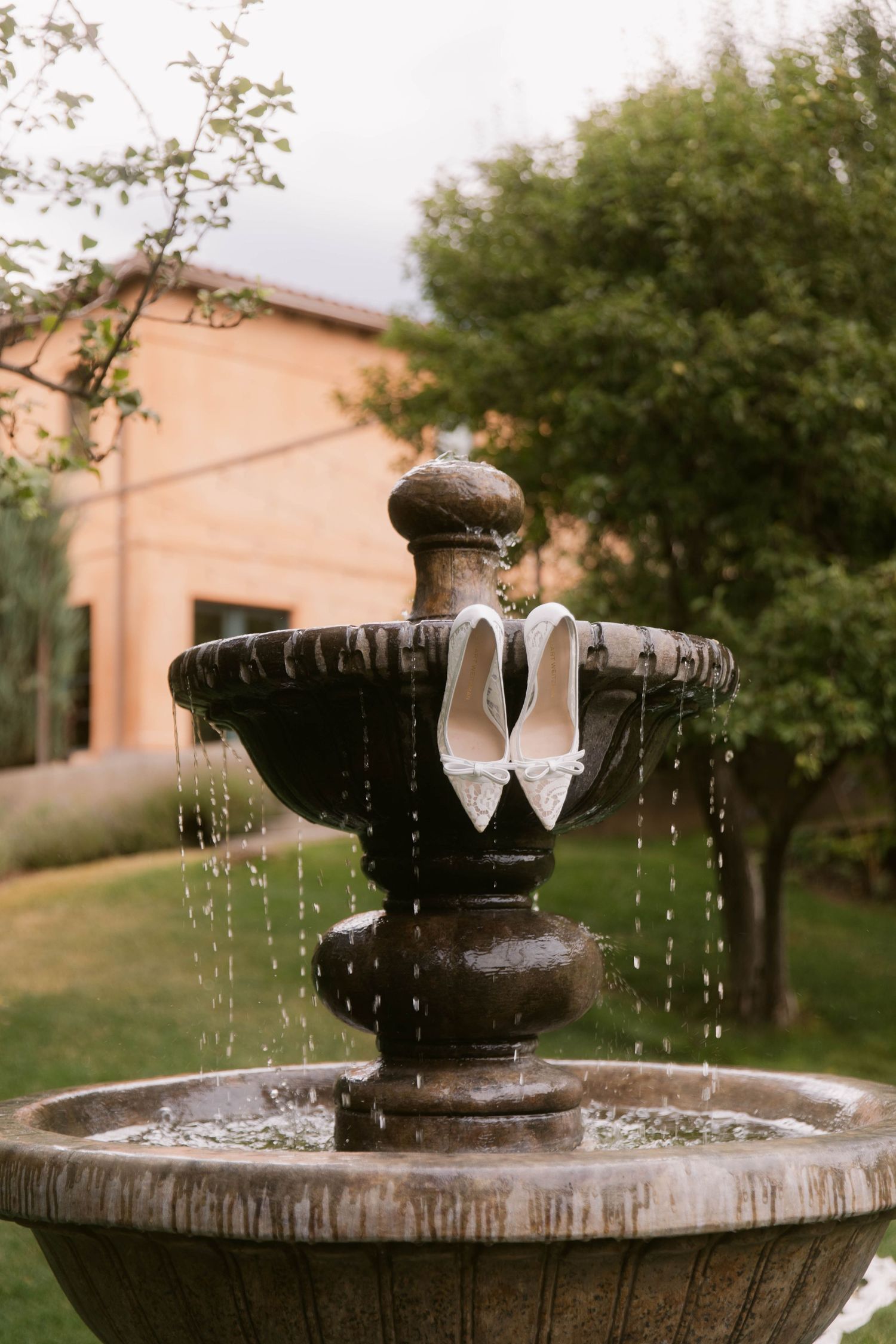 White bridal heels displayed on a rustic stone garden fountain, with lush greenery and a terracotta building in the background.