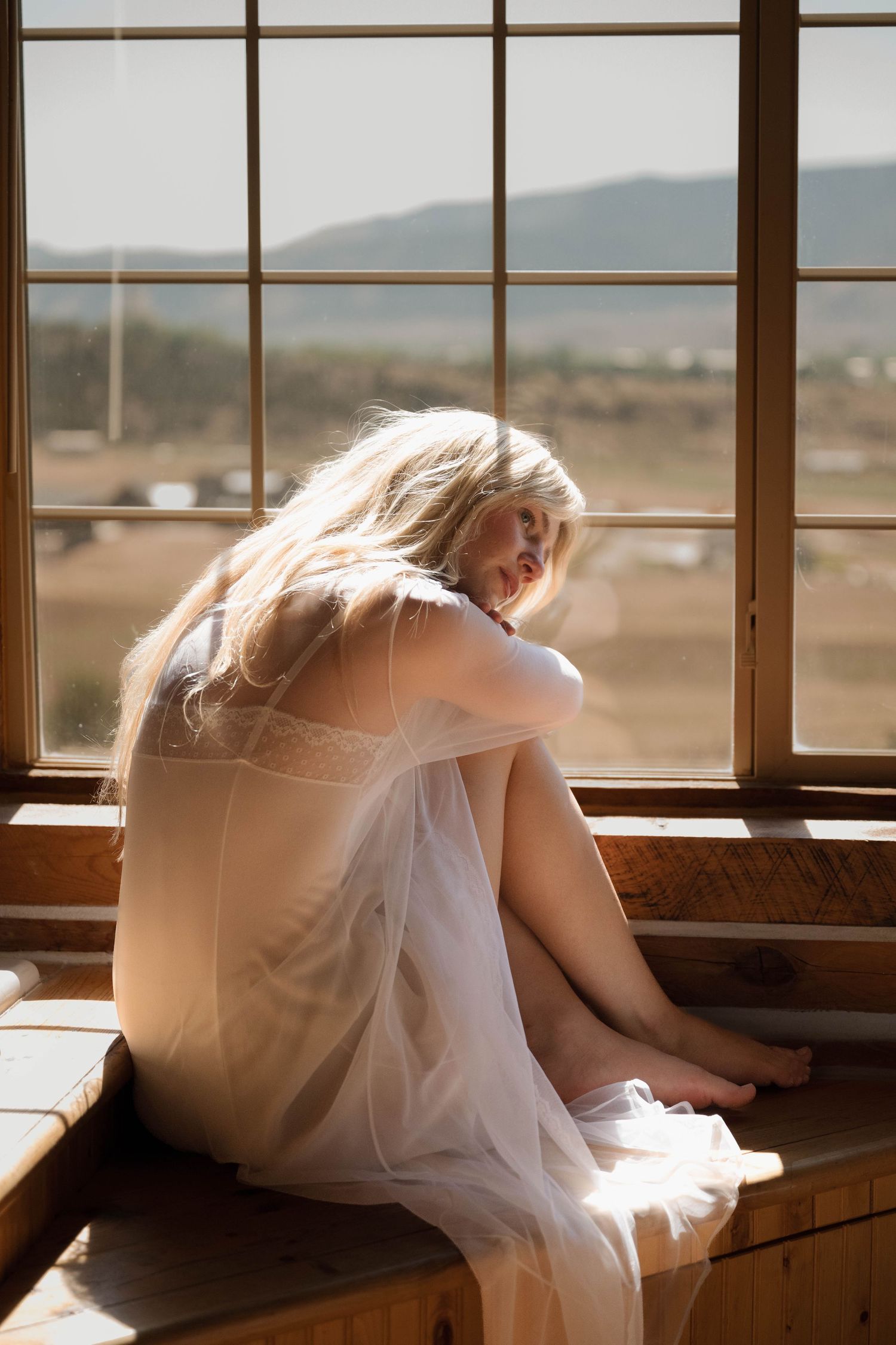 Blonde woman in sheer white dress sitting curled up on wooden window bench looking down contemplatively.