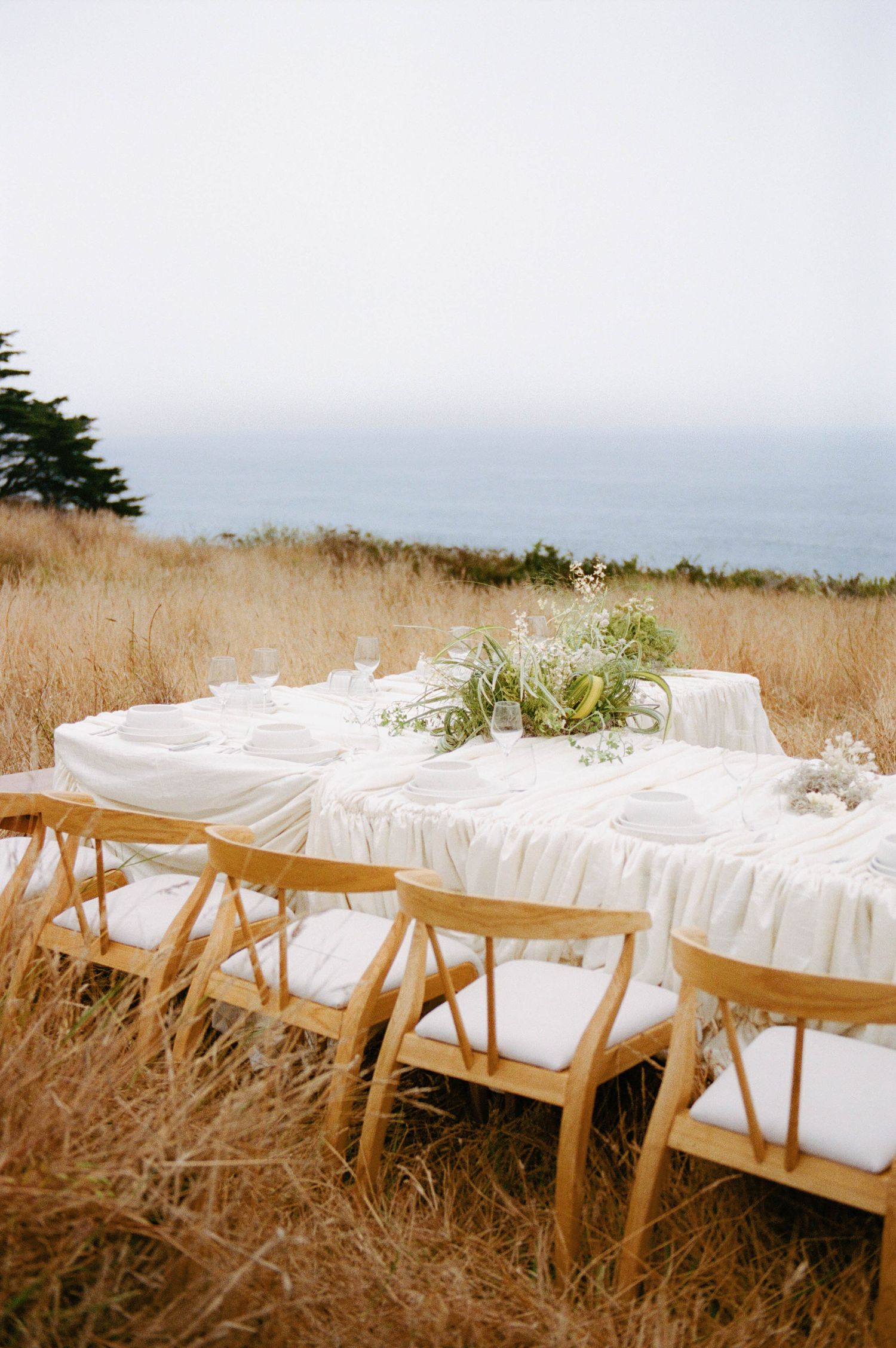 Elegant outdoor dining table with white linen and floral centerpiece set in golden grass overlooking the ocean.