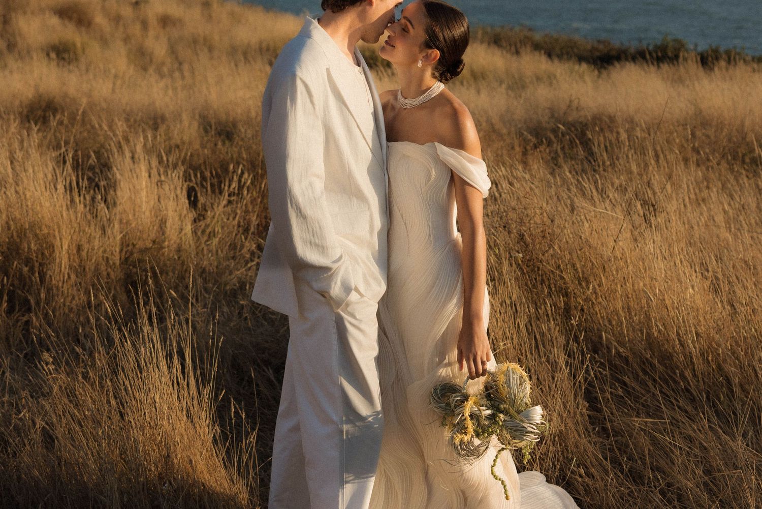 Bride and groom embrace in golden field, bride wearing off-shoulder white gown holding bouquet at sunset.