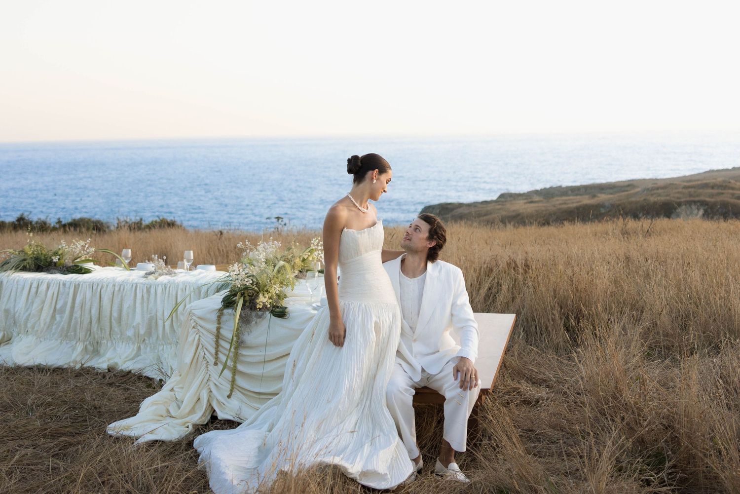 Bride in white gown stands beside groom in white suit at outdoor coastal wedding table with ocean views.