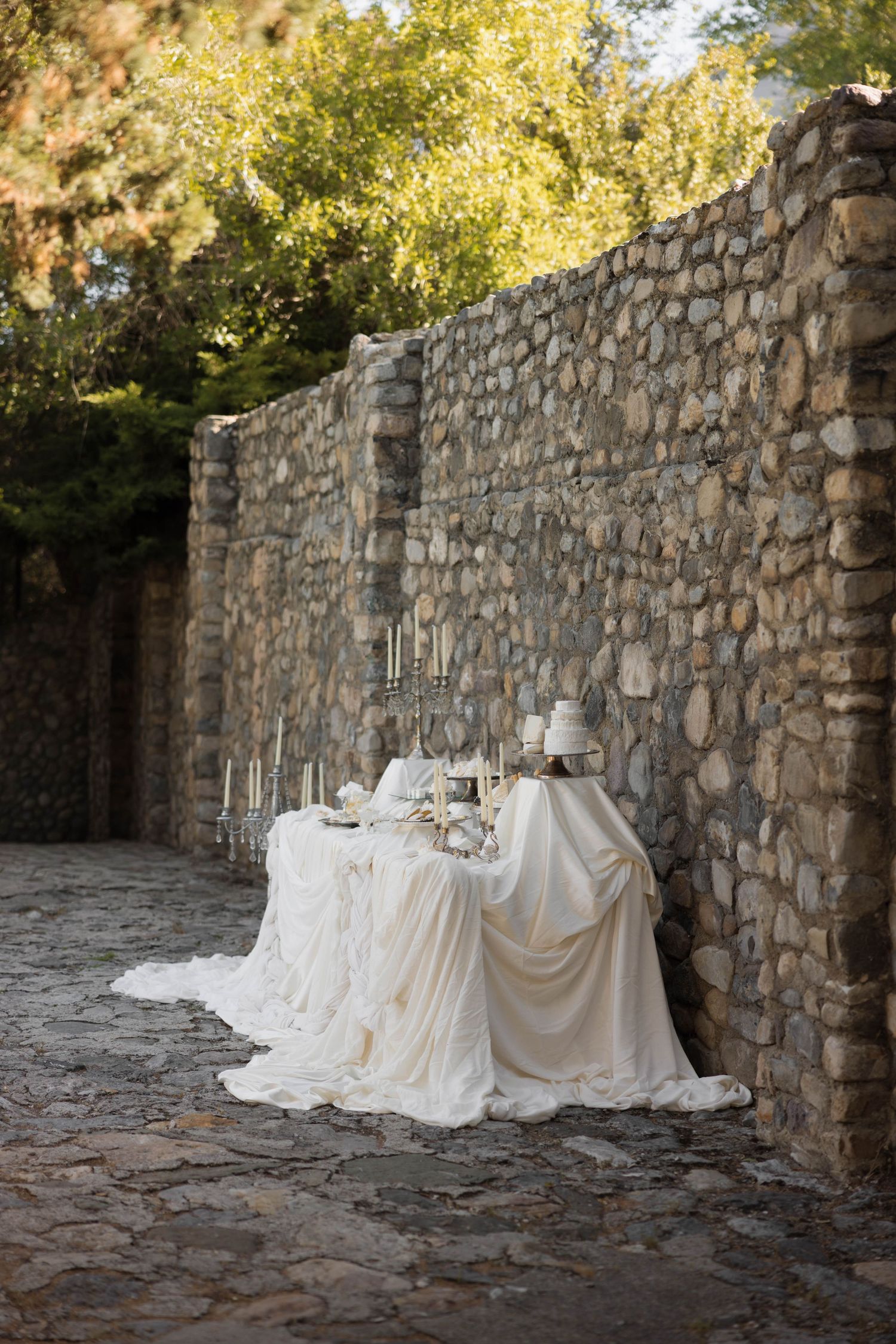 Elegant outdoor wedding table draped in white fabric against a rustic stone wall, adorned with candles and a cake.