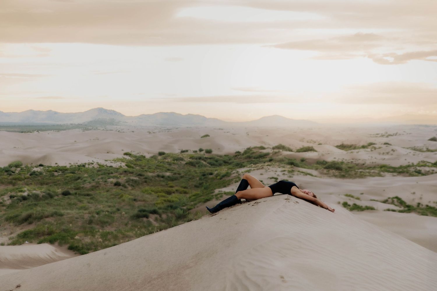Person lying on sandy desert dune at sunset with mountains and sparse greenery background.