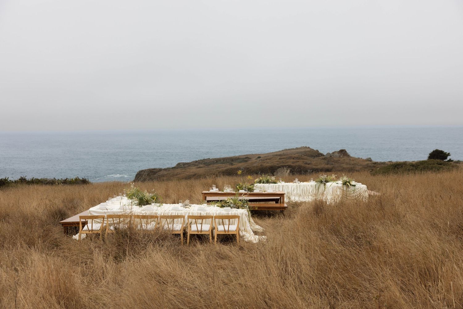 Outdoor dining table set in golden coastal grasslands with ocean and rocky cliffs in the misty background.