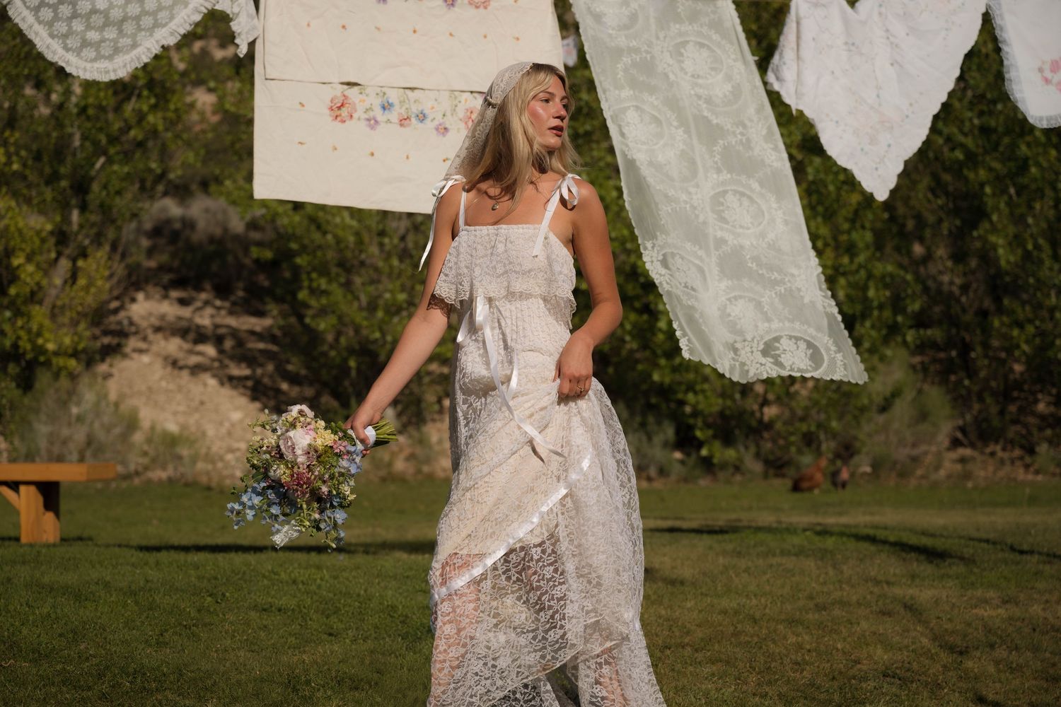 Bride in flowing lace wedding dress holding wildflower bouquet standing near hanging vintage linens outdoors.