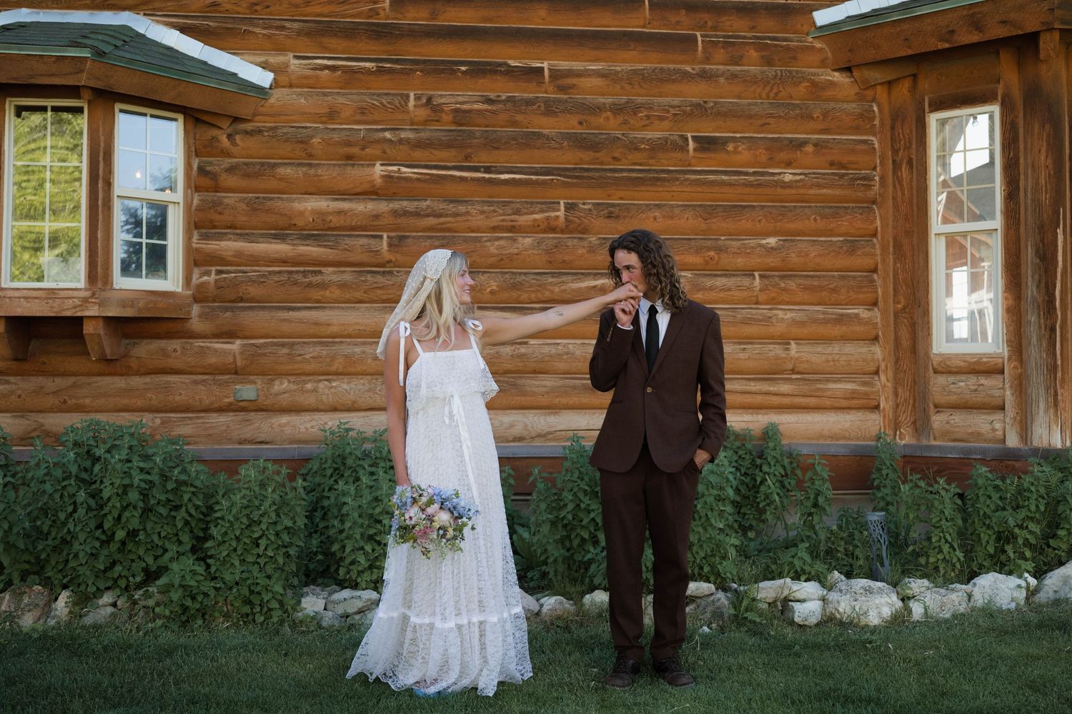 Bride in white boho dress and groom in brown suit holding hands outside rustic log cabin venue.