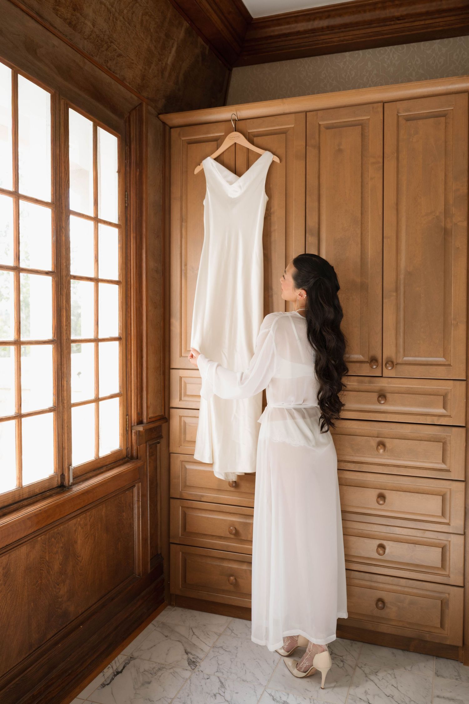 Bride in white robe admires her wedding gown hanging on a wooden wardrobe in an elegant dressing room.