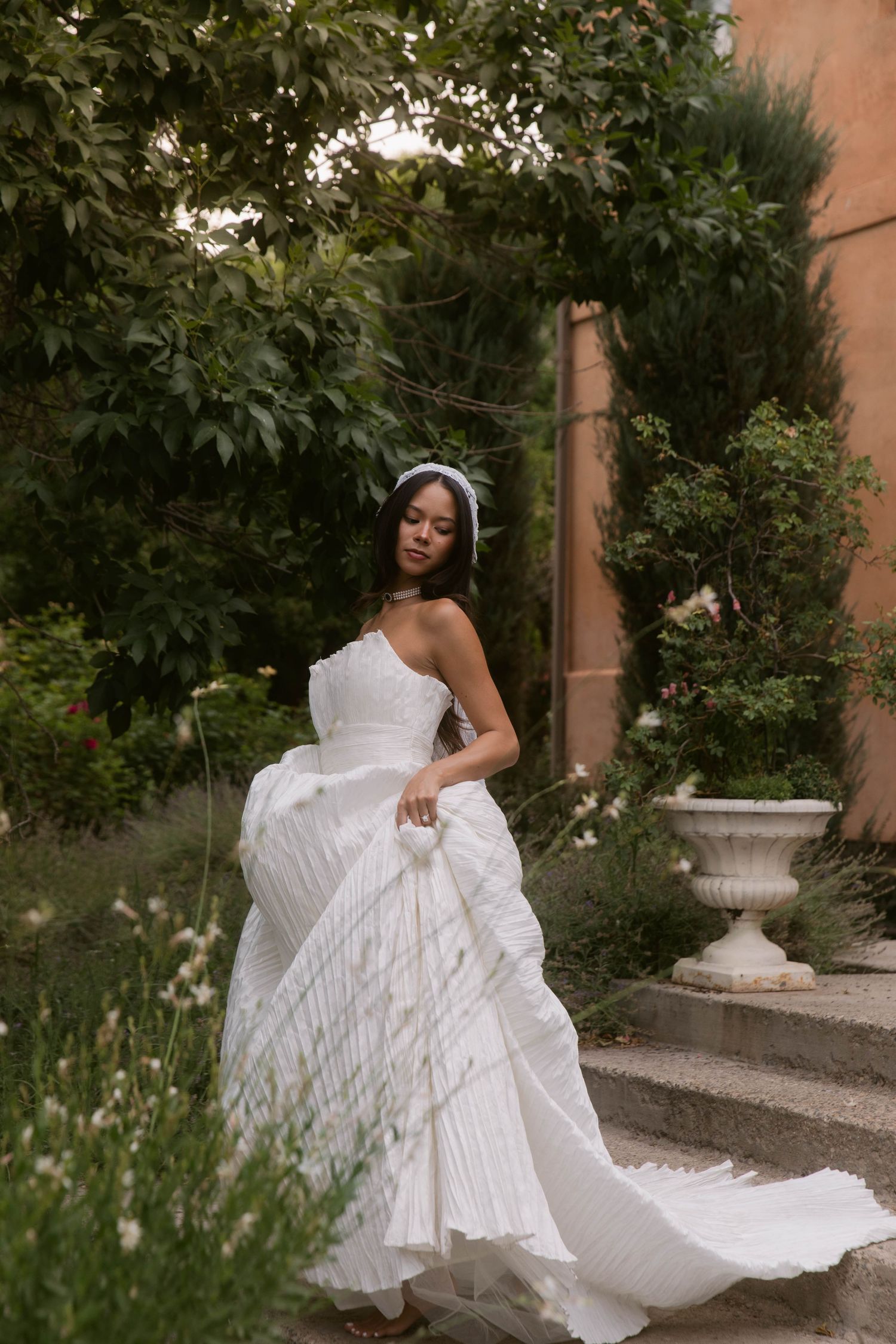 Bride in strapless white pleated ballgown with veil poses on garden steps surrounded by lush greenery.