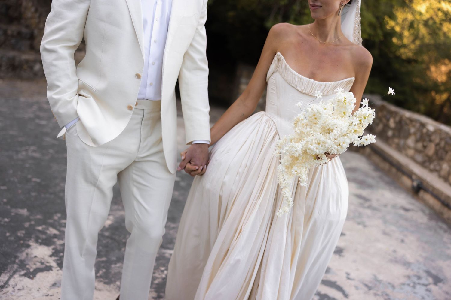 Bride and groom holding hands in elegant white wedding attire, bride carrying a white floral bouquet outdoors.