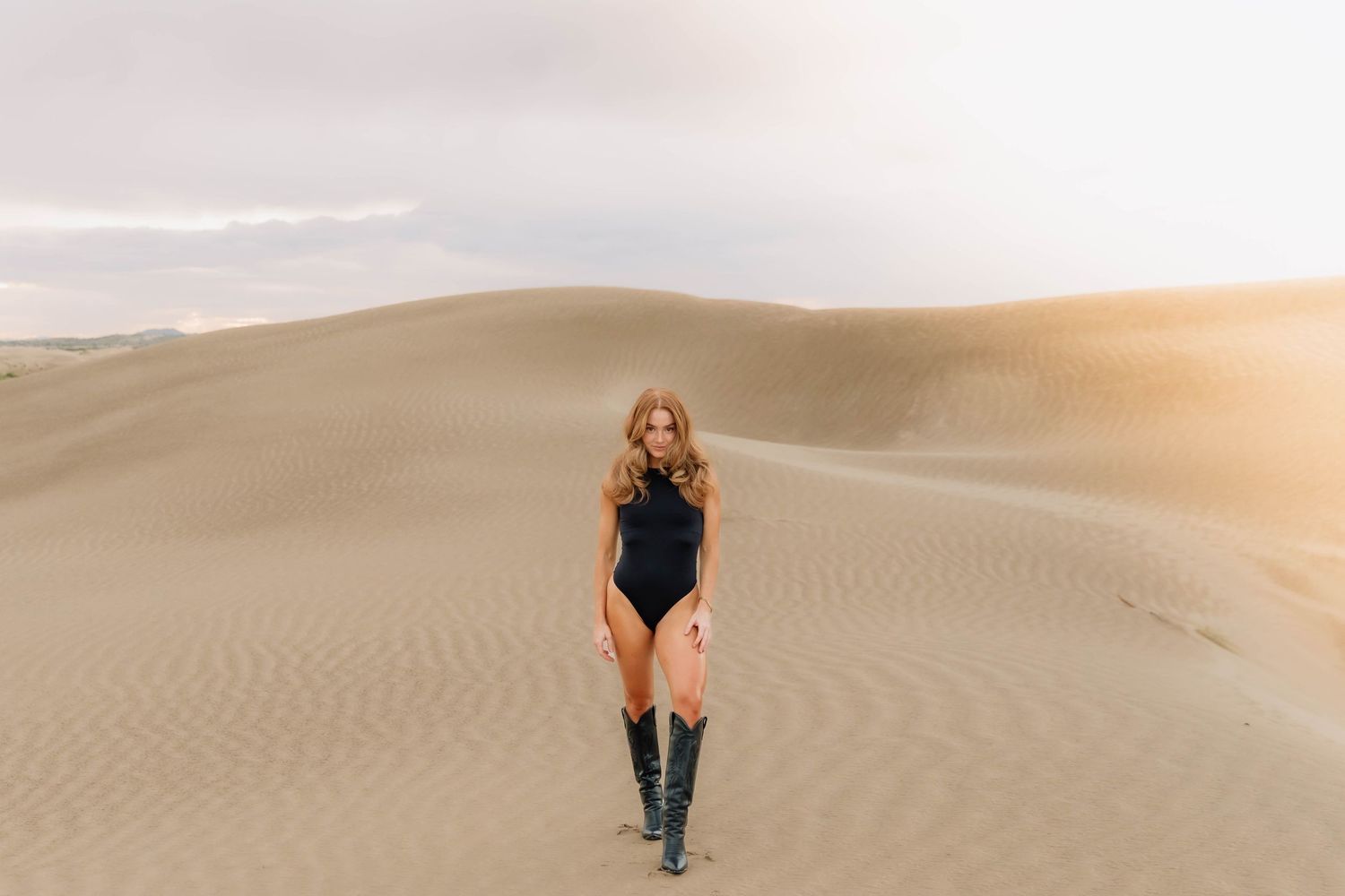 Blonde woman in black bodysuit and knee-high boots standing on vast sandy desert dunes at sunset.