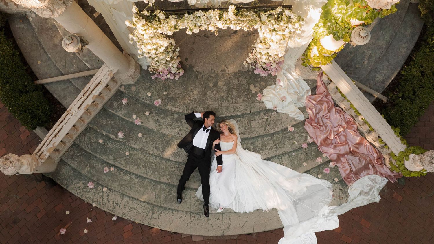 Aerial view of bride and groom lying on stone steps surrounded by pink and white floral decor.
