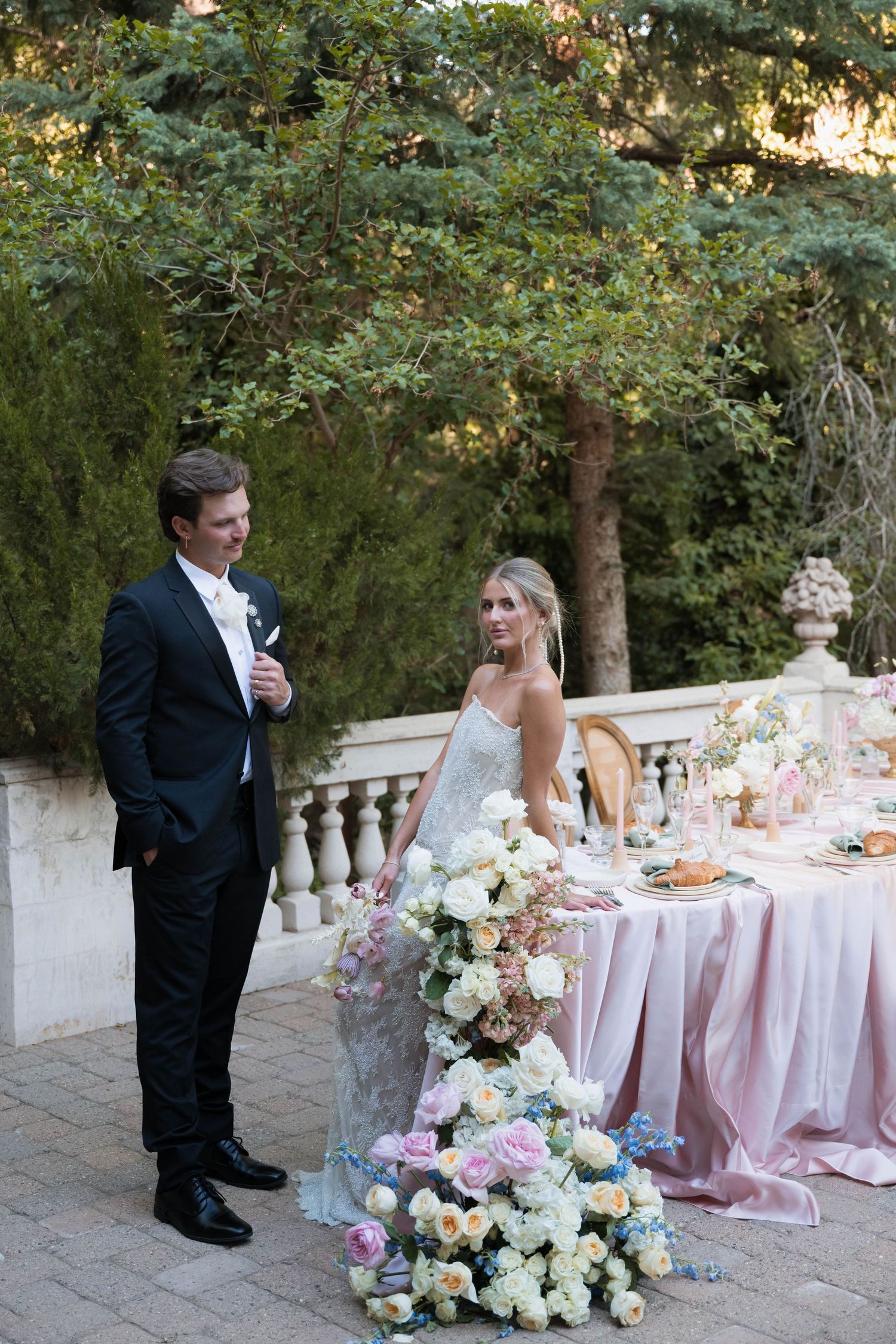 Bride and groom stand by an elegantly decorated outdoor reception table with cascading floral arrangements.