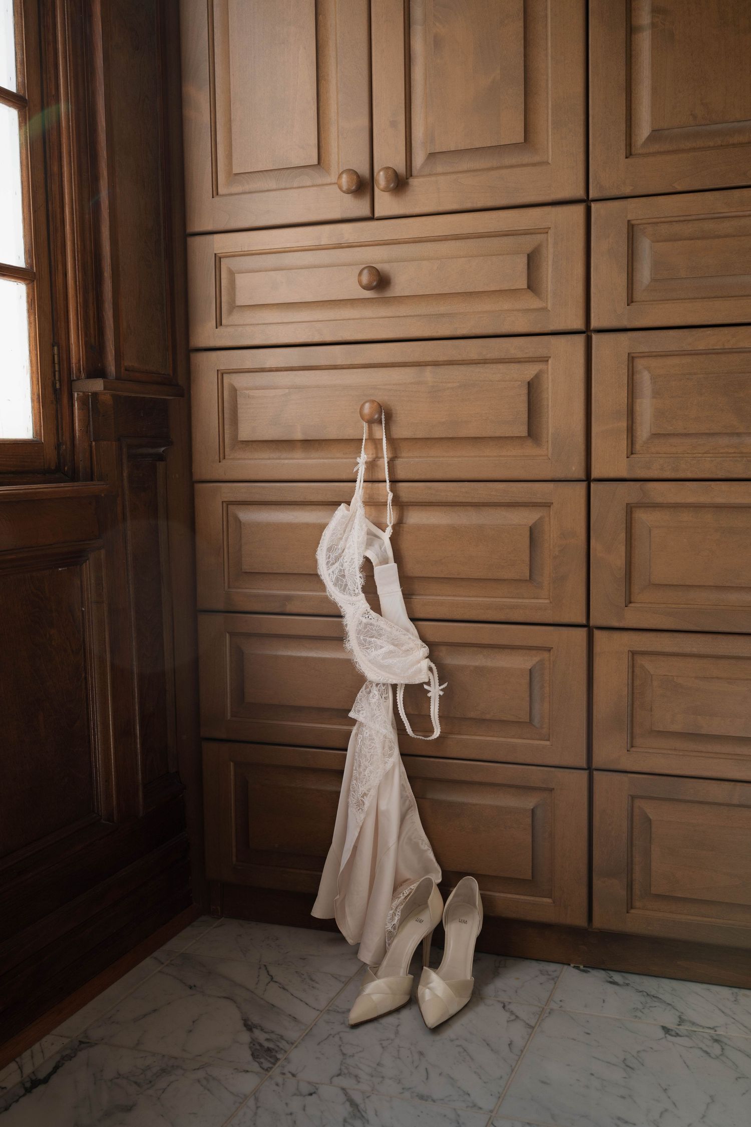 Ivory bridal lingerie and heels hanging and placed against elegant wooden wardrobe in a marble dressing room.