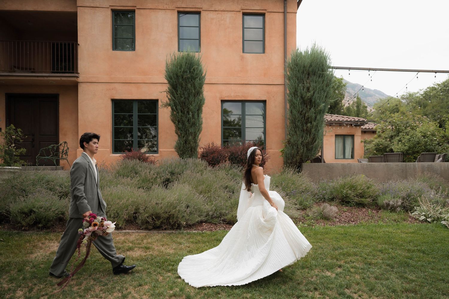 A bride and groom walk across a green lawn beside a terracotta villa with cypress trees and mountain views.