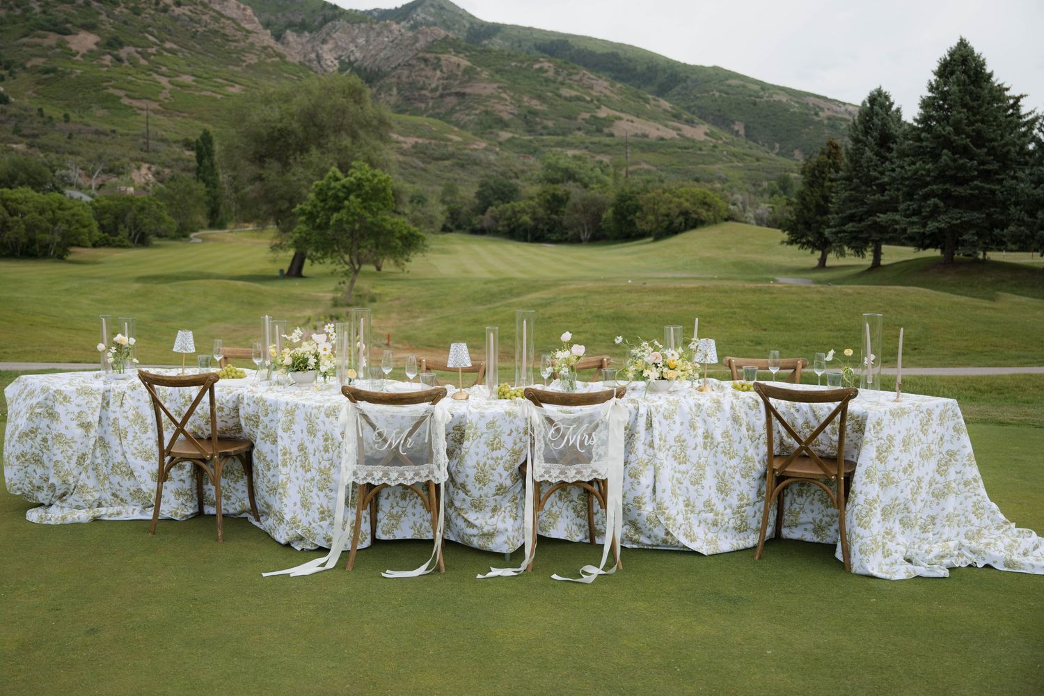 Elegant outdoor wedding reception table set on a golf course with floral linens, wooden chairs, and mountain backdrop.