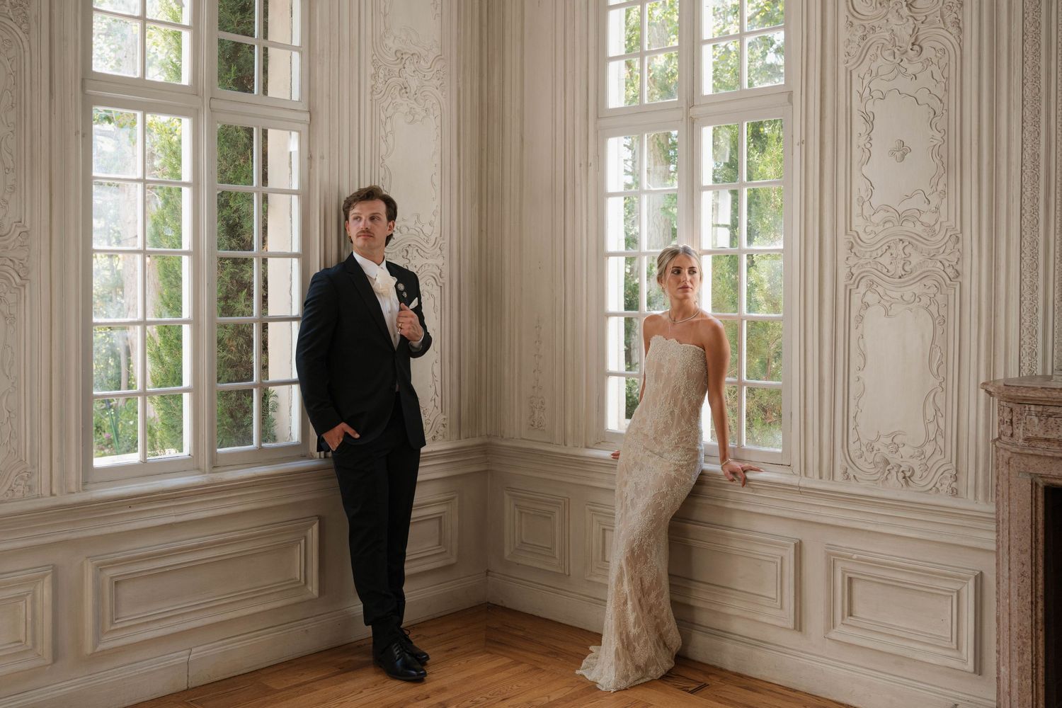 Elegant bride and groom pose separately by tall ornate white French windows in a grand ballroom.