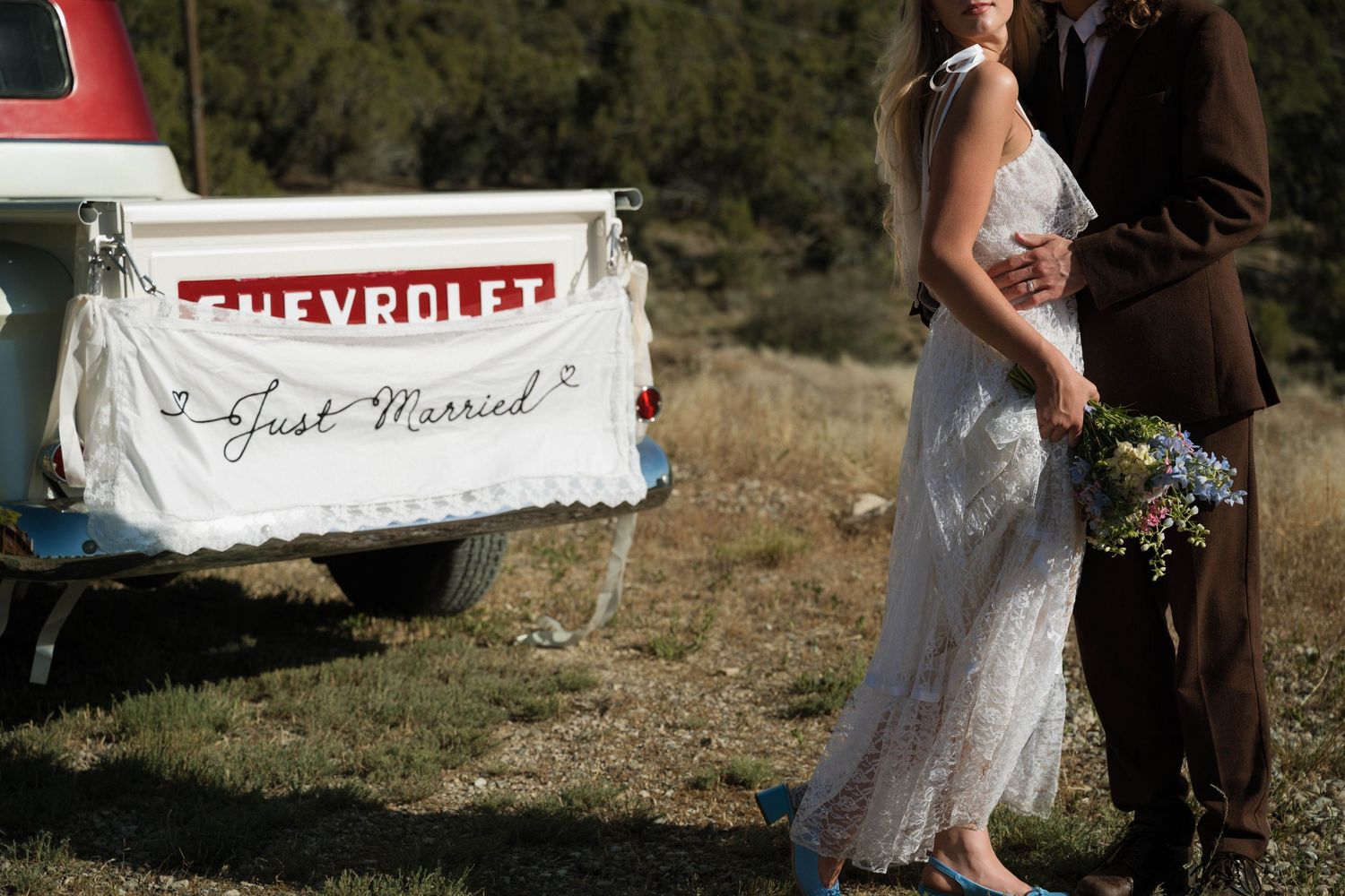 Newlyweds embrace beside vintage Chevrolet truck with Just Married banner in scenic outdoor setting.