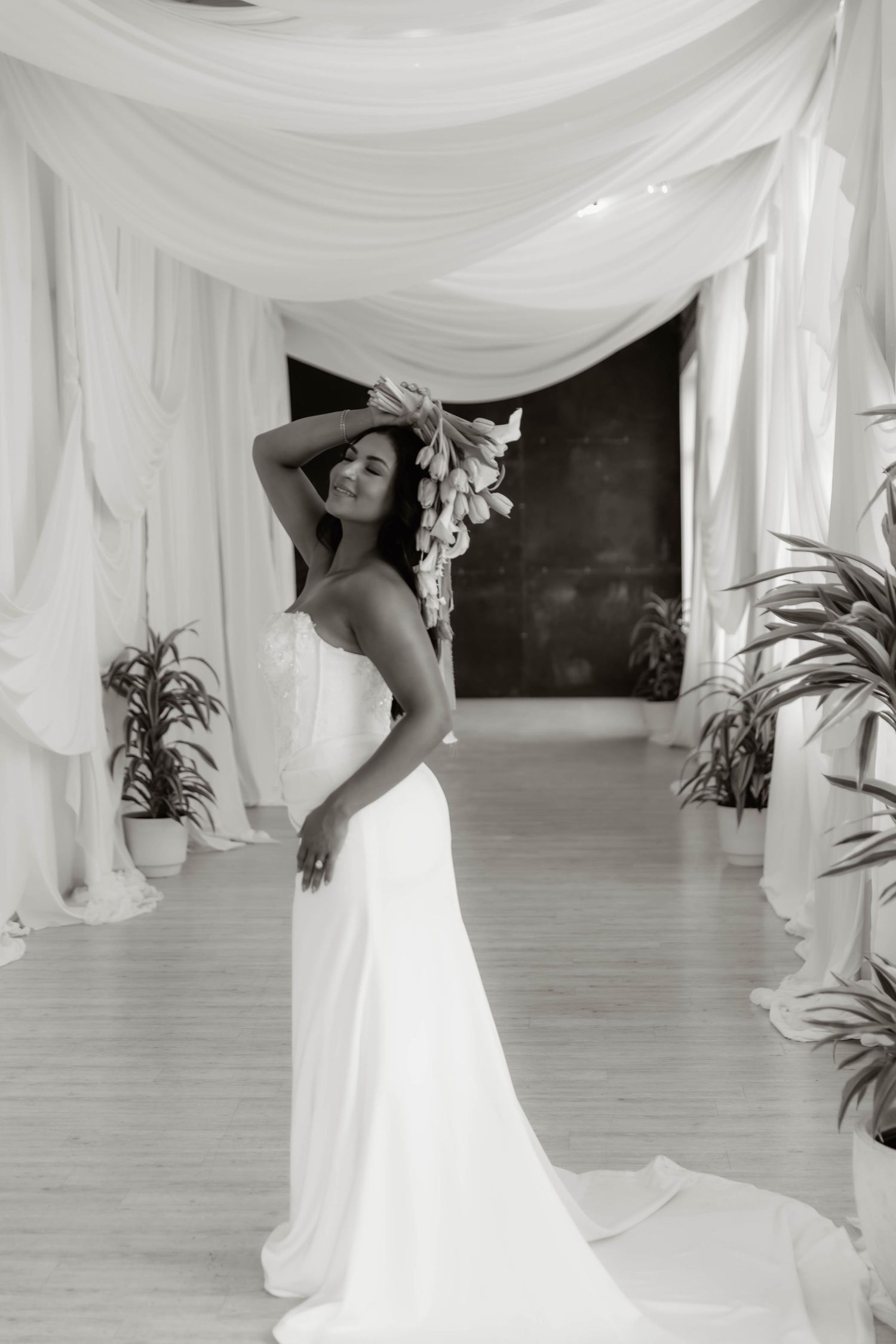 Elegant bride in strapless white gown holding flowers poses under draped white fabric archway, black and white photo.