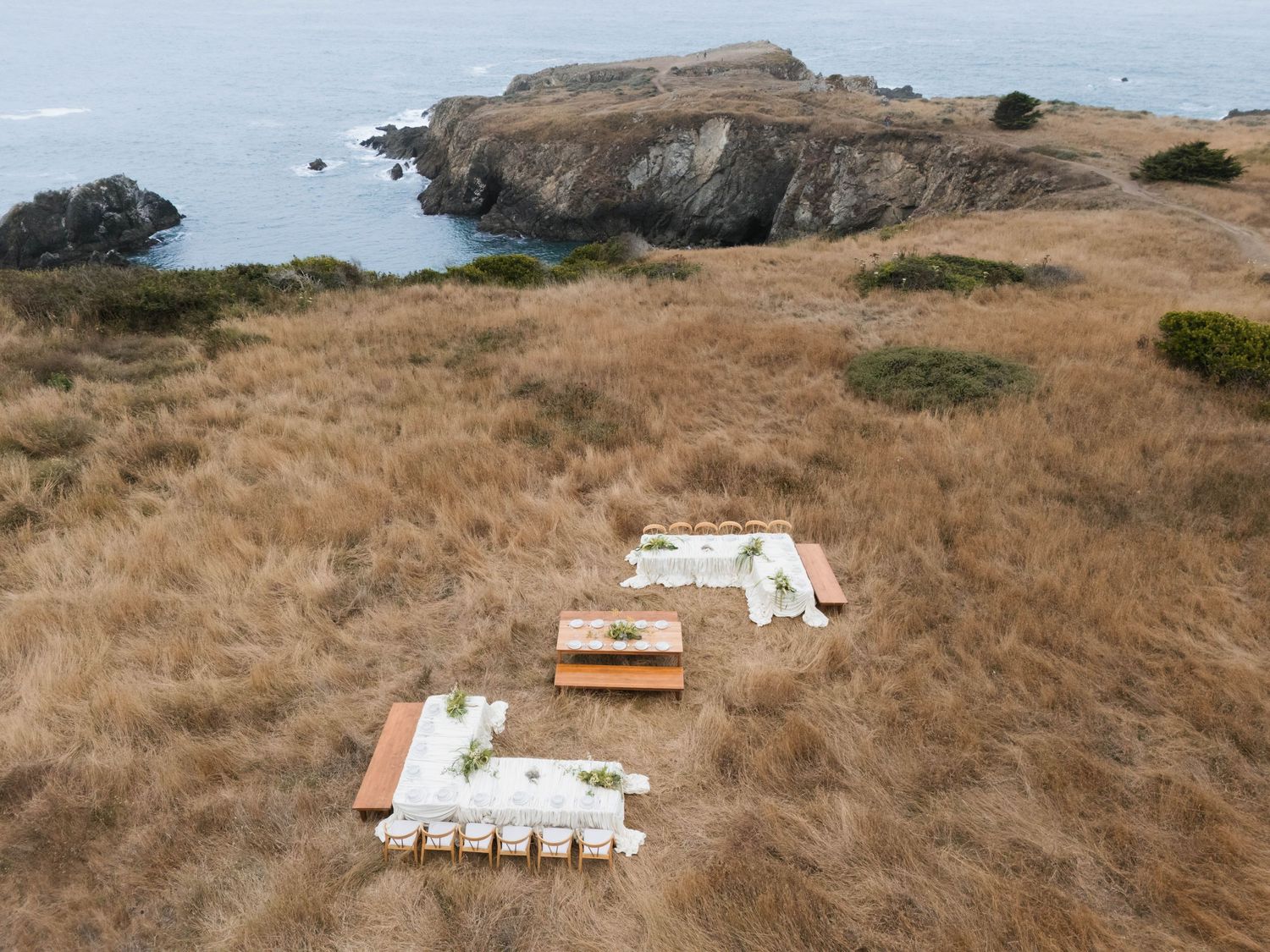 Aerial view of rustic wooden furniture arrangements on golden coastal hillside with rocky ocean shoreline in background.