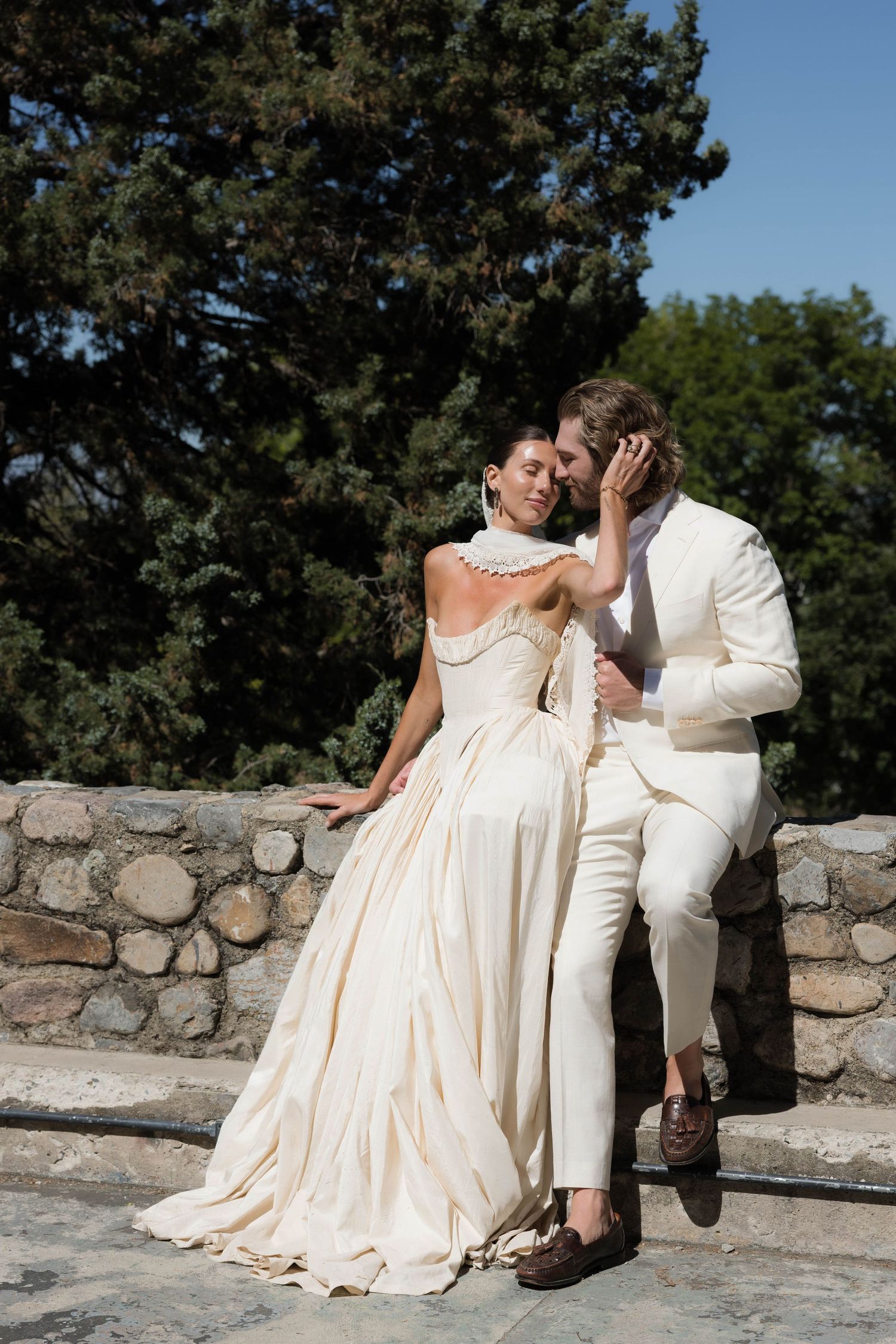 Elegant bride in ivory gown and groom in white suit share romantic moment on stone terrace with pine trees.