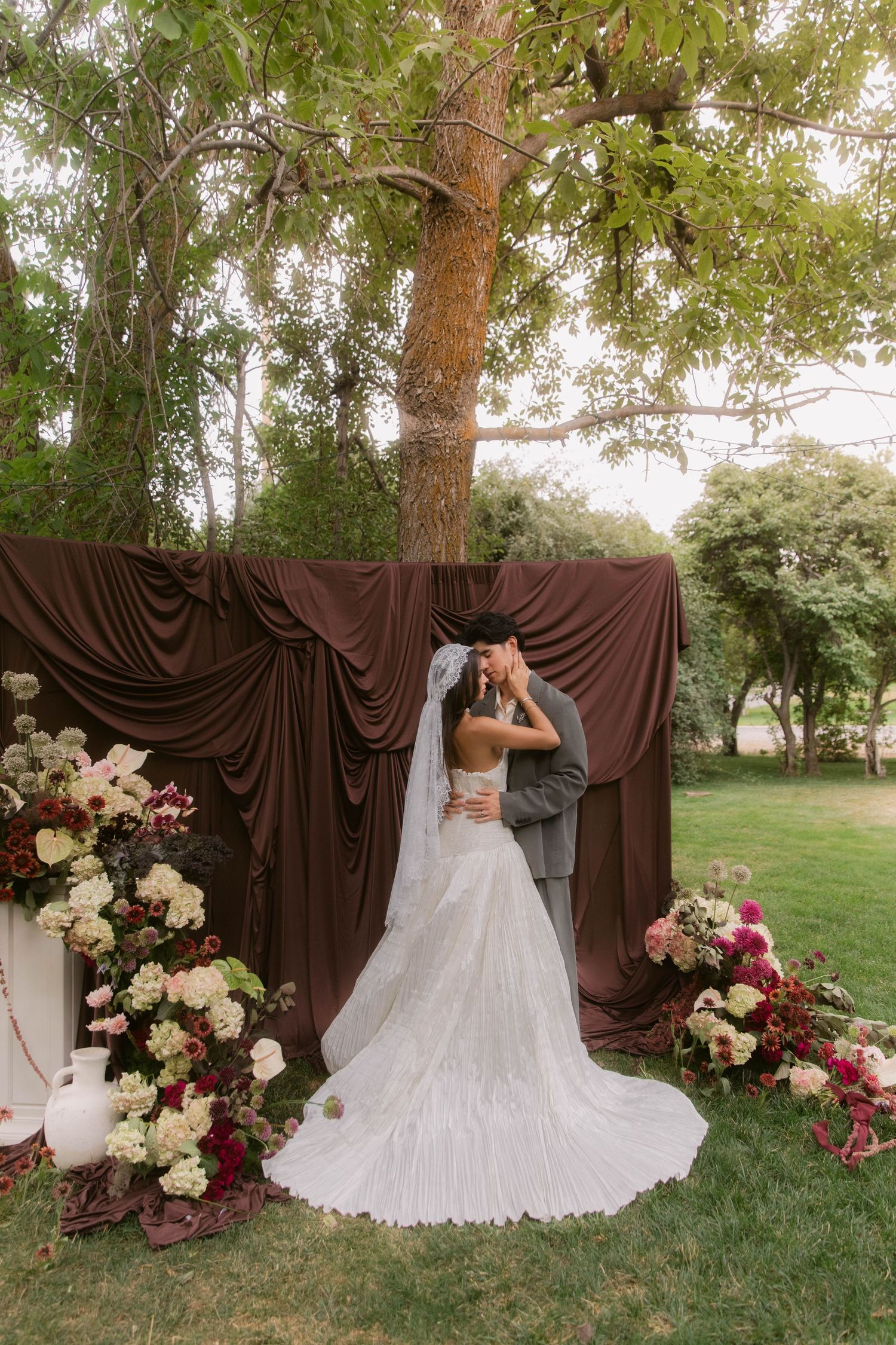 Bride and groom share a kiss under a tree with brown draping and floral arrangements at an outdoor wedding ceremony.