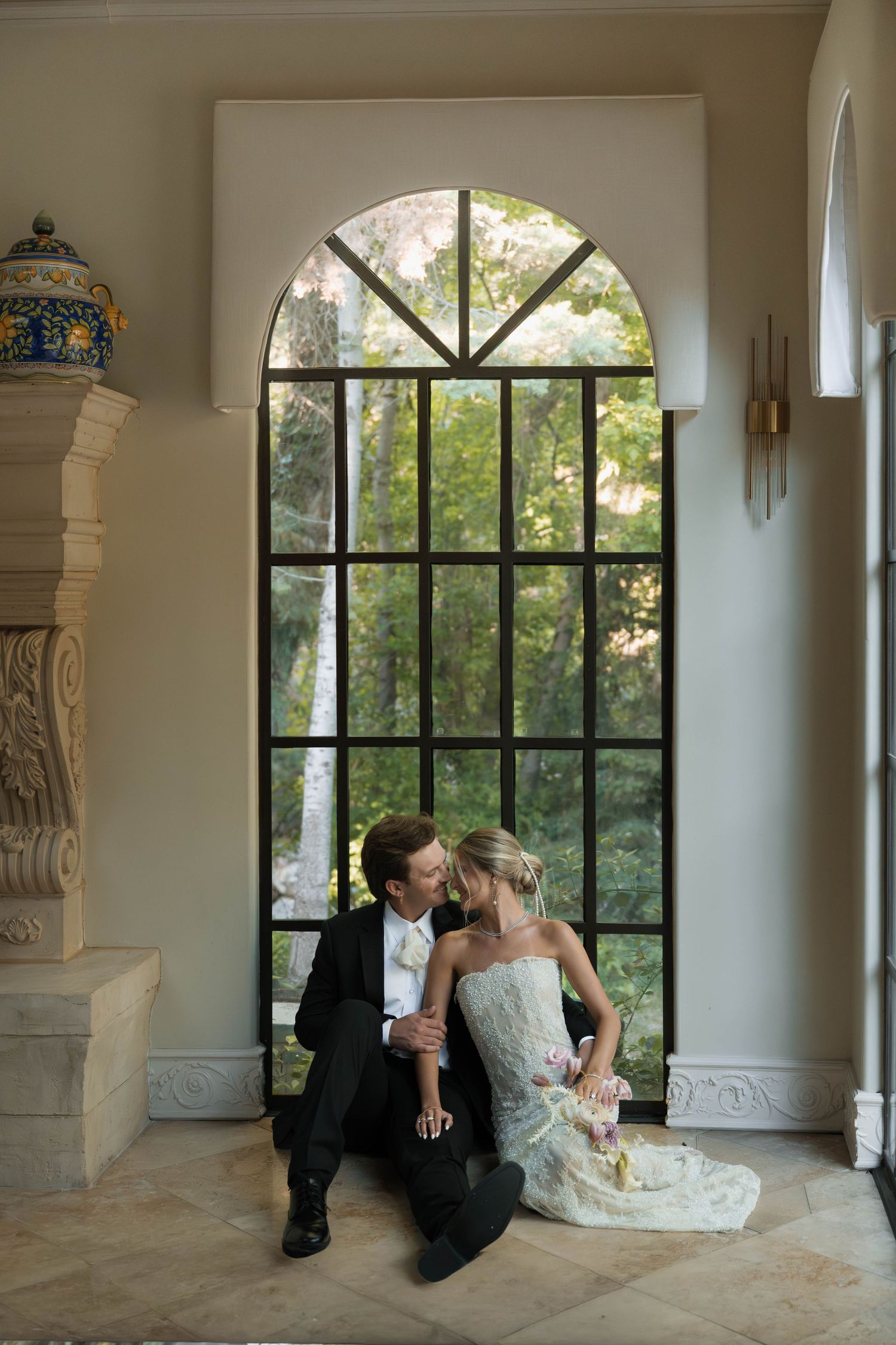 Bride and groom sit together on the floor in front of a large arched window in an elegant venue.