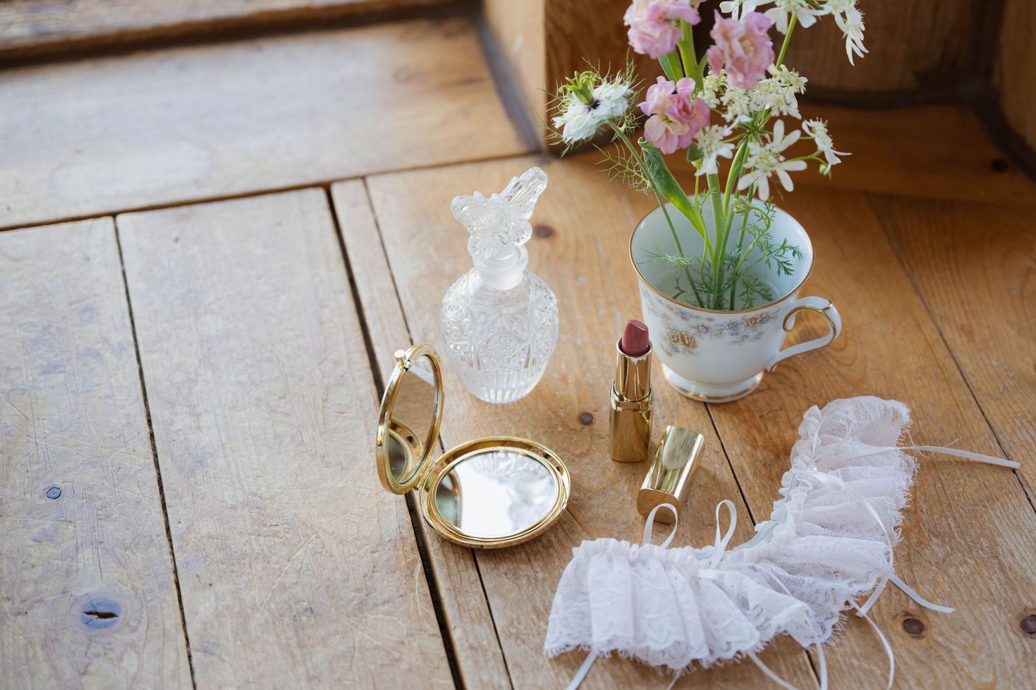 Vintage wedding flat lay with gold compact mirror, lipstick, perfume bottle, lace garter, and floral teacup on rustic wood floor.