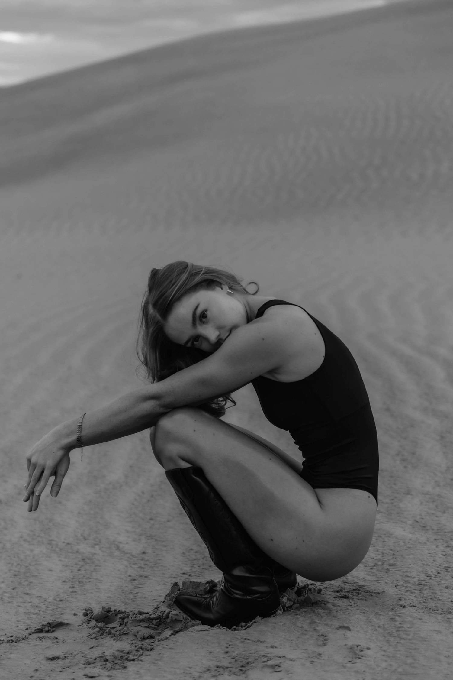 Black and white photo of woman in black bodysuit sitting on sand dunes in desert landscape.