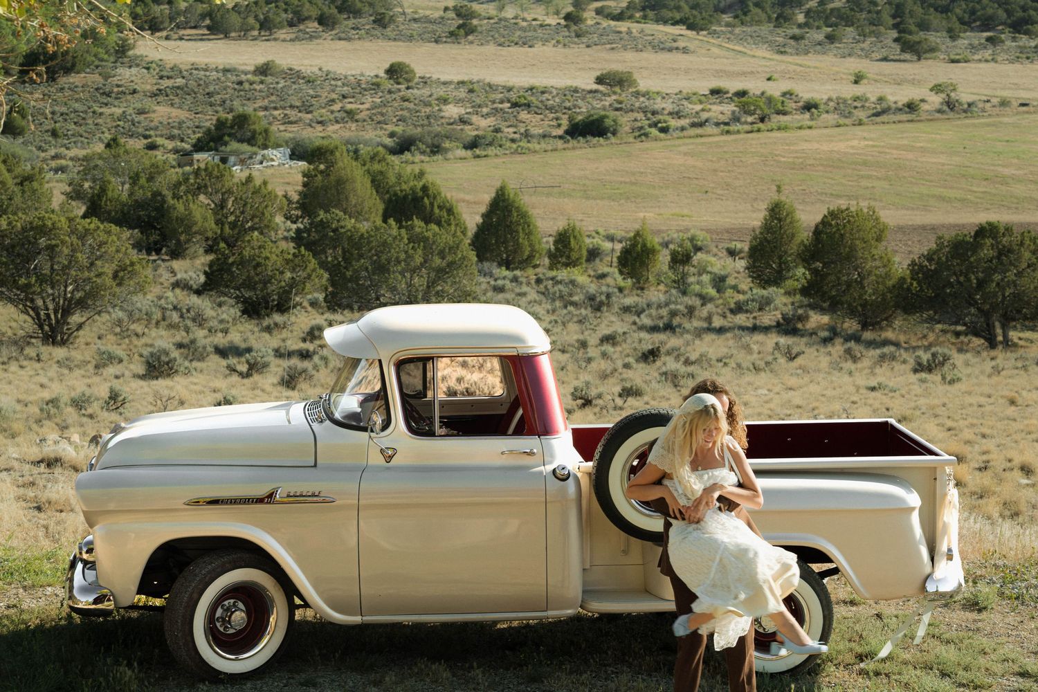 Bride in white gown sitting on a vintage cream Chevrolet pickup truck in a scenic rural countryside setting.