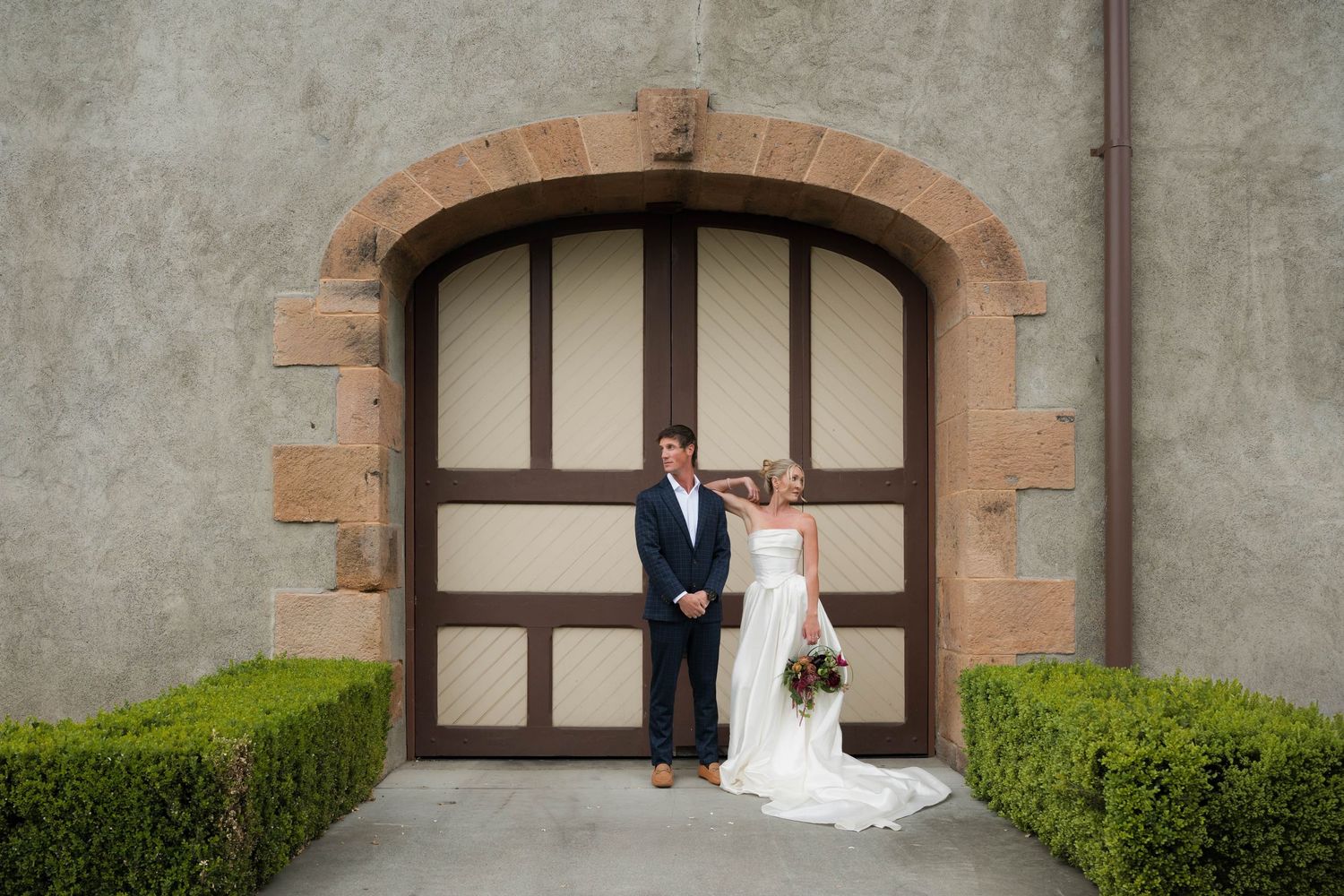 Bride and groom standing before large arched wooden doors at elegant stone venue on their wedding day.