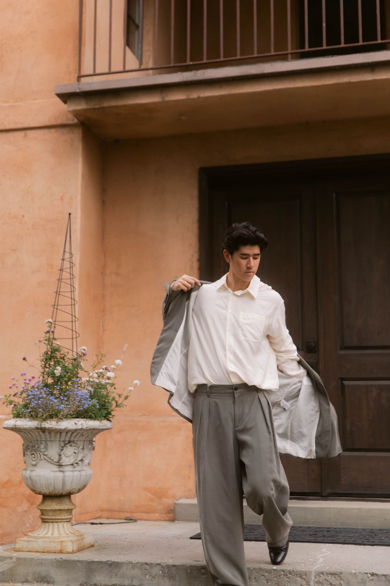 Young man in white shirt and gray trousers putting on jacket outside rustic Mediterranean building with flower pot.