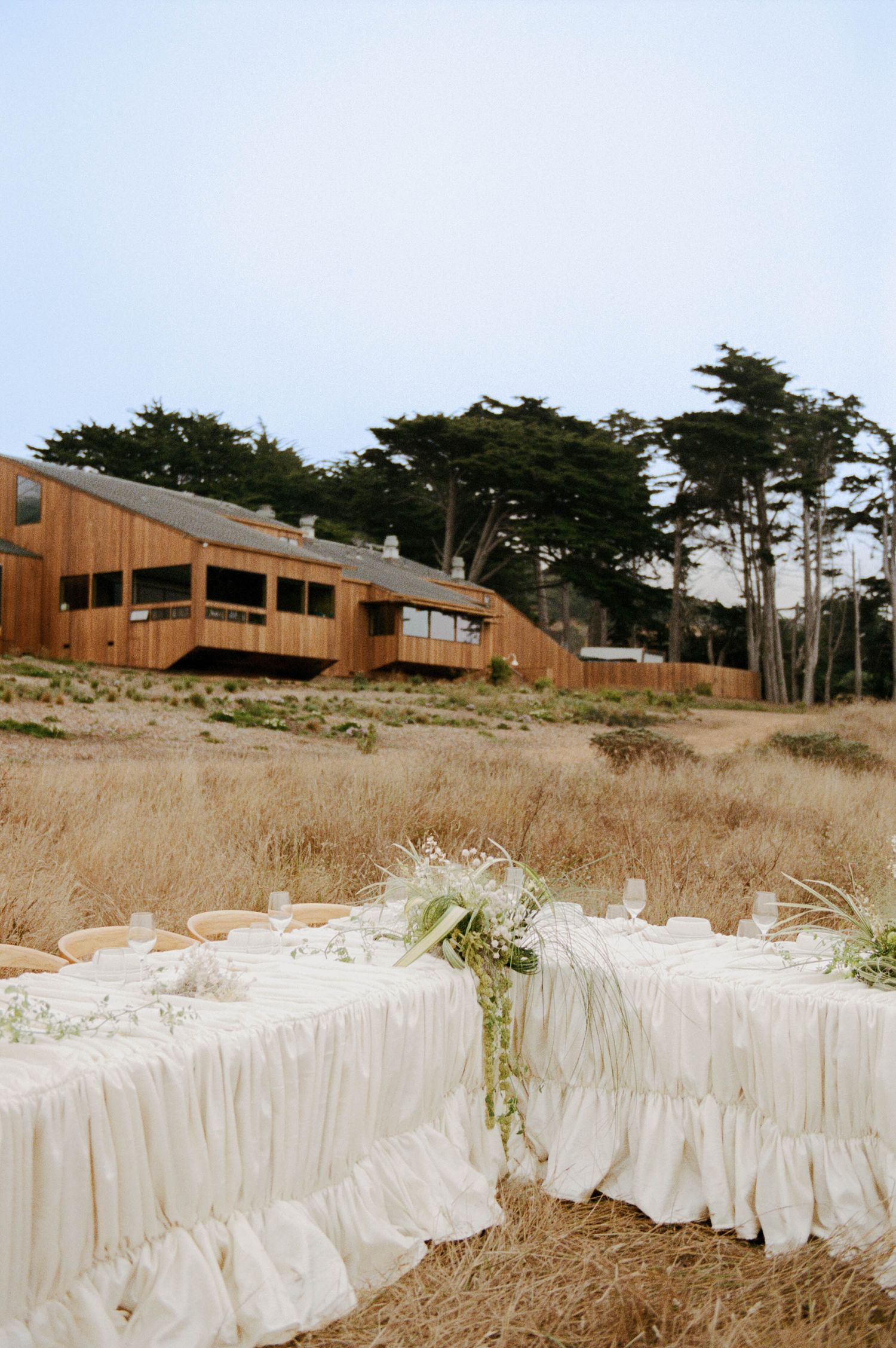 Outdoor wedding reception table with white ruffled linens set in golden coastal meadow before modern wood building.
