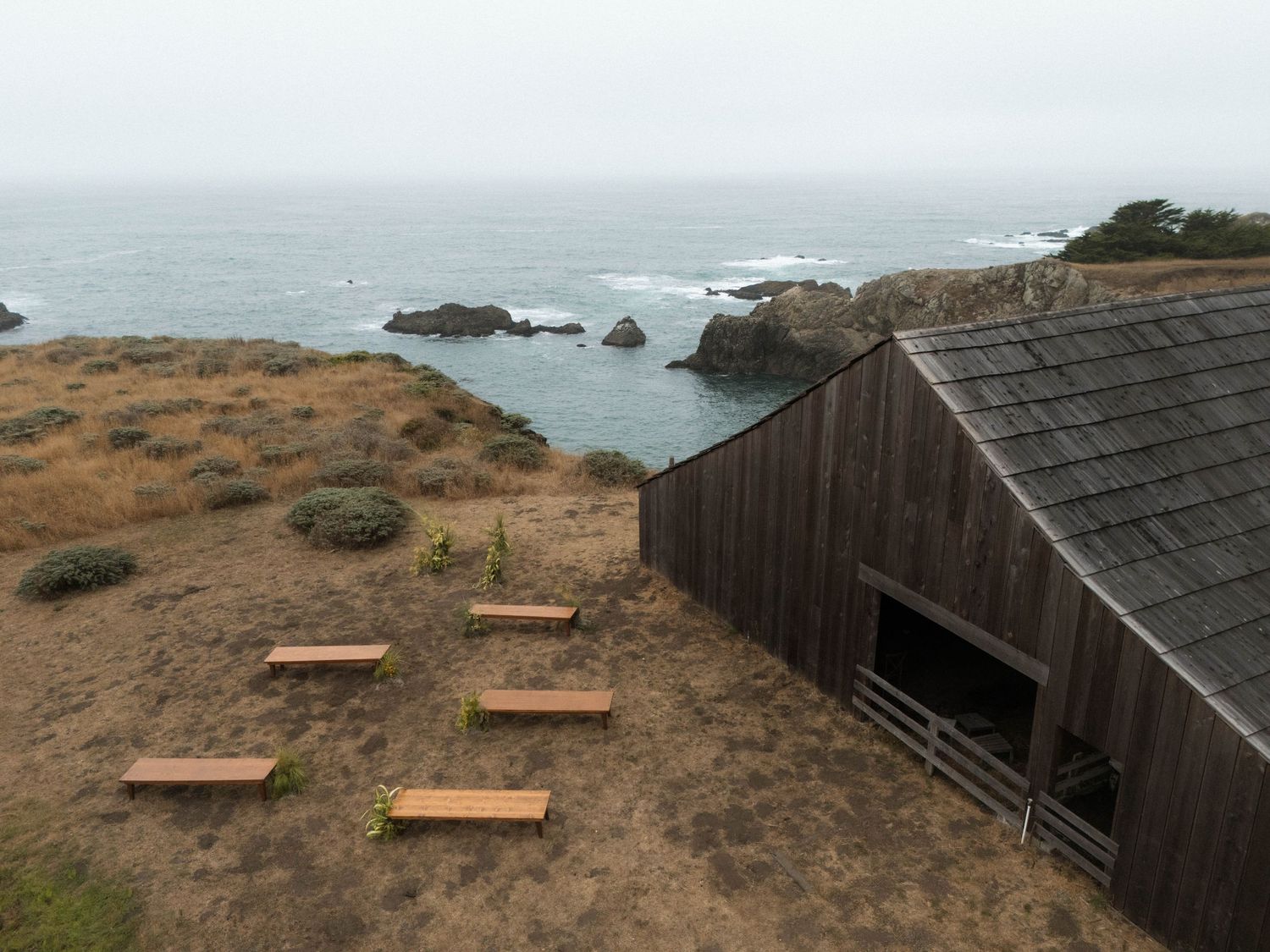 Rustic wooden barn on a coastal bluff overlooking rocky Pacific Ocean shoreline with wooden benches on dry grass.