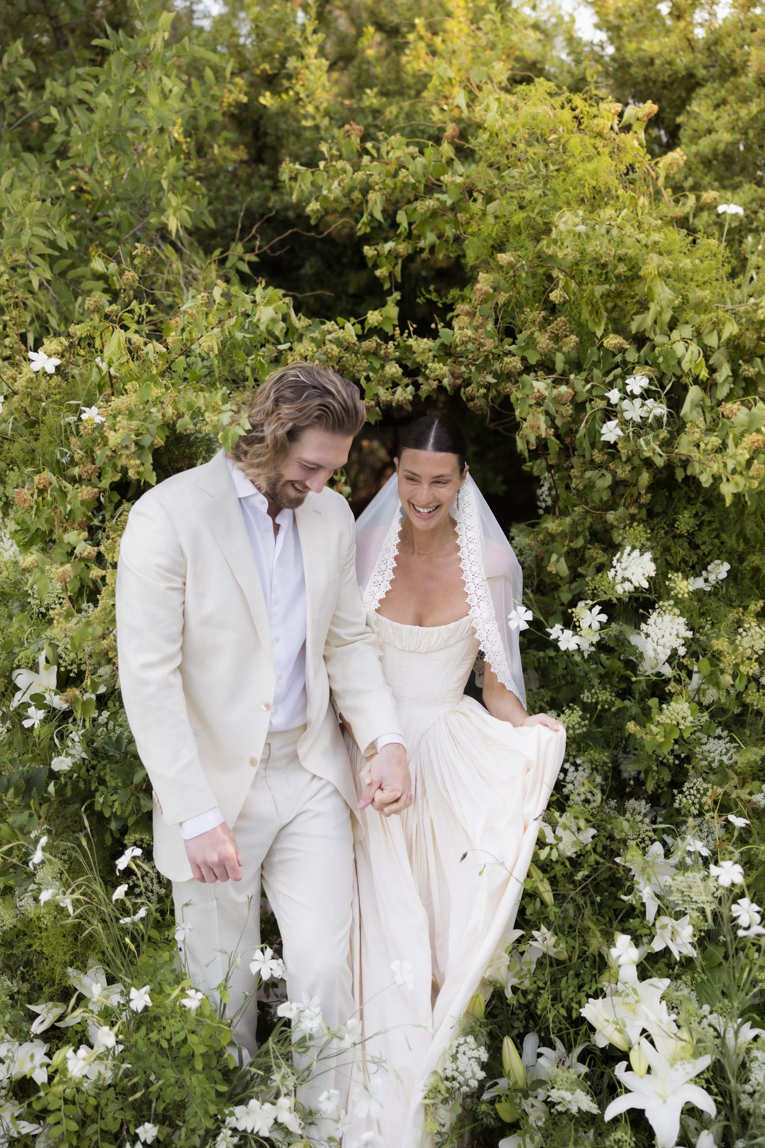 Bride and groom walk through lush garden archway, she in white gown and veil, he in cream suit.