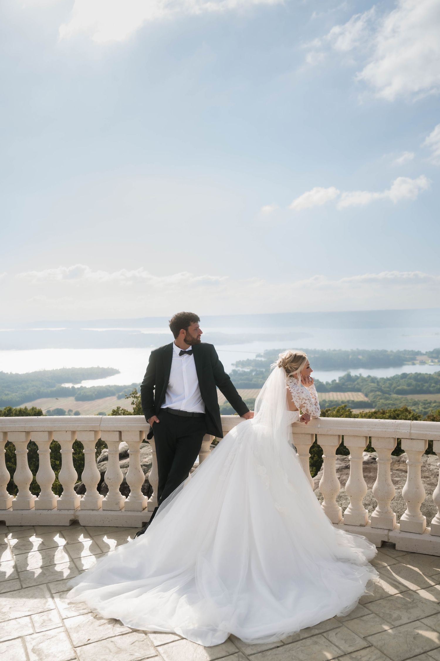 Bride in white gown and groom in tuxedo pose on elegant balcony overlooking scenic coastal landscape.