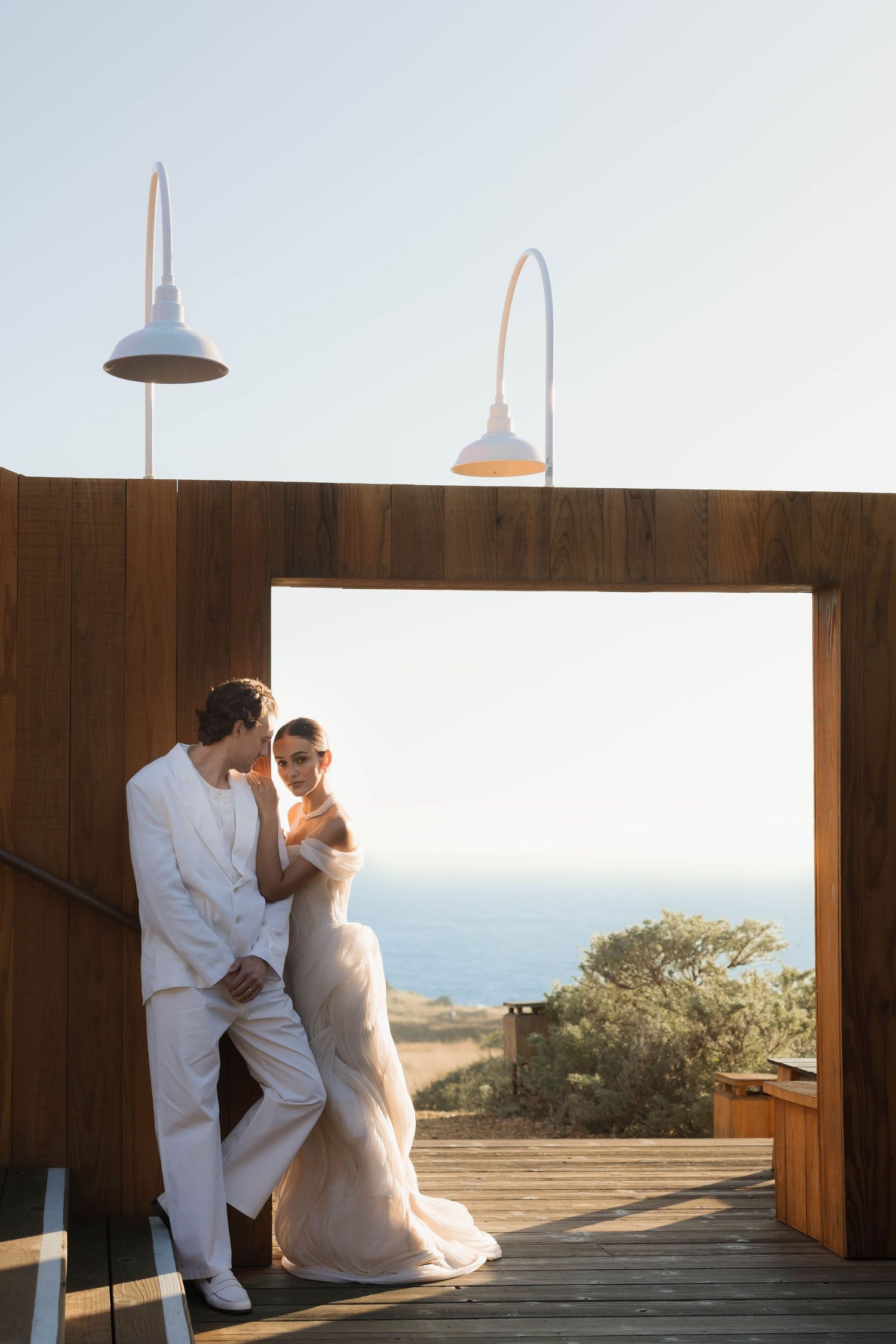 Bride and groom in elegant attire embrace on a wooden deck overlooking the ocean at golden hour.