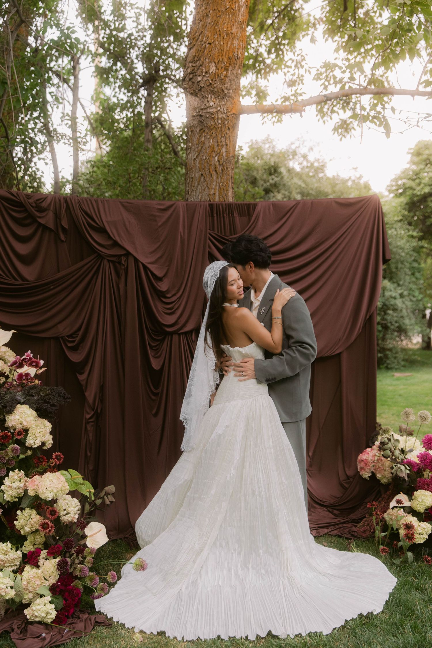 Bride and groom embrace before draped brown fabric backdrop with floral arrangements at outdoor wedding ceremony.