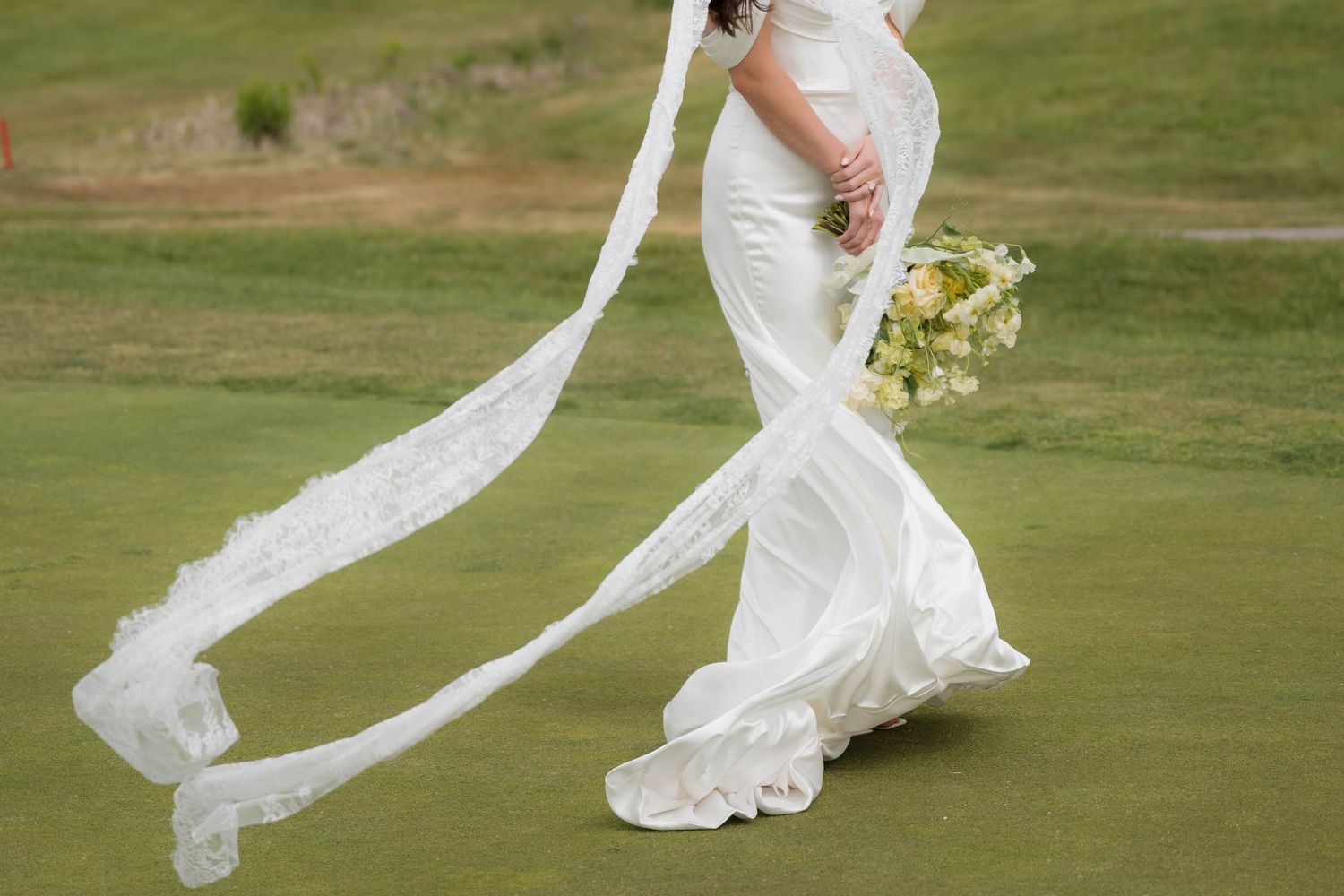 Bride in elegant fitted white wedding dress with flowing lace veil walks across lush green golf course.