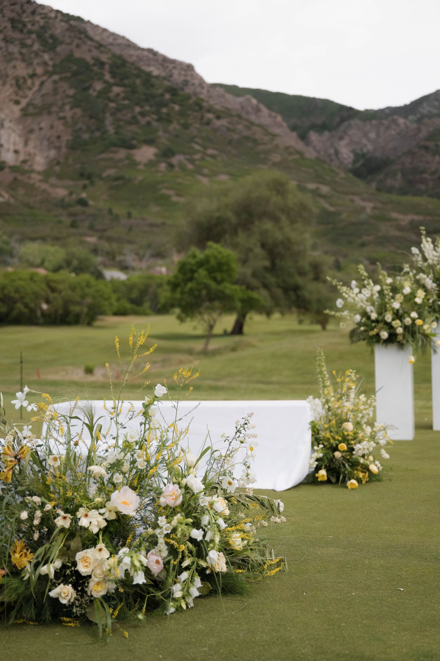 Close-up of lush white and yellow wildflower wedding arrangements on green grass with mountain view.