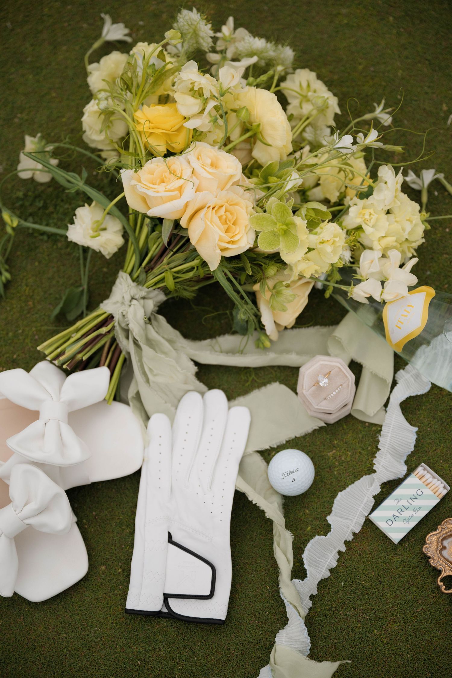 Flat lay of wedding details including yellow bouquet, white golf gloves, golf ball, and ribbon on green.