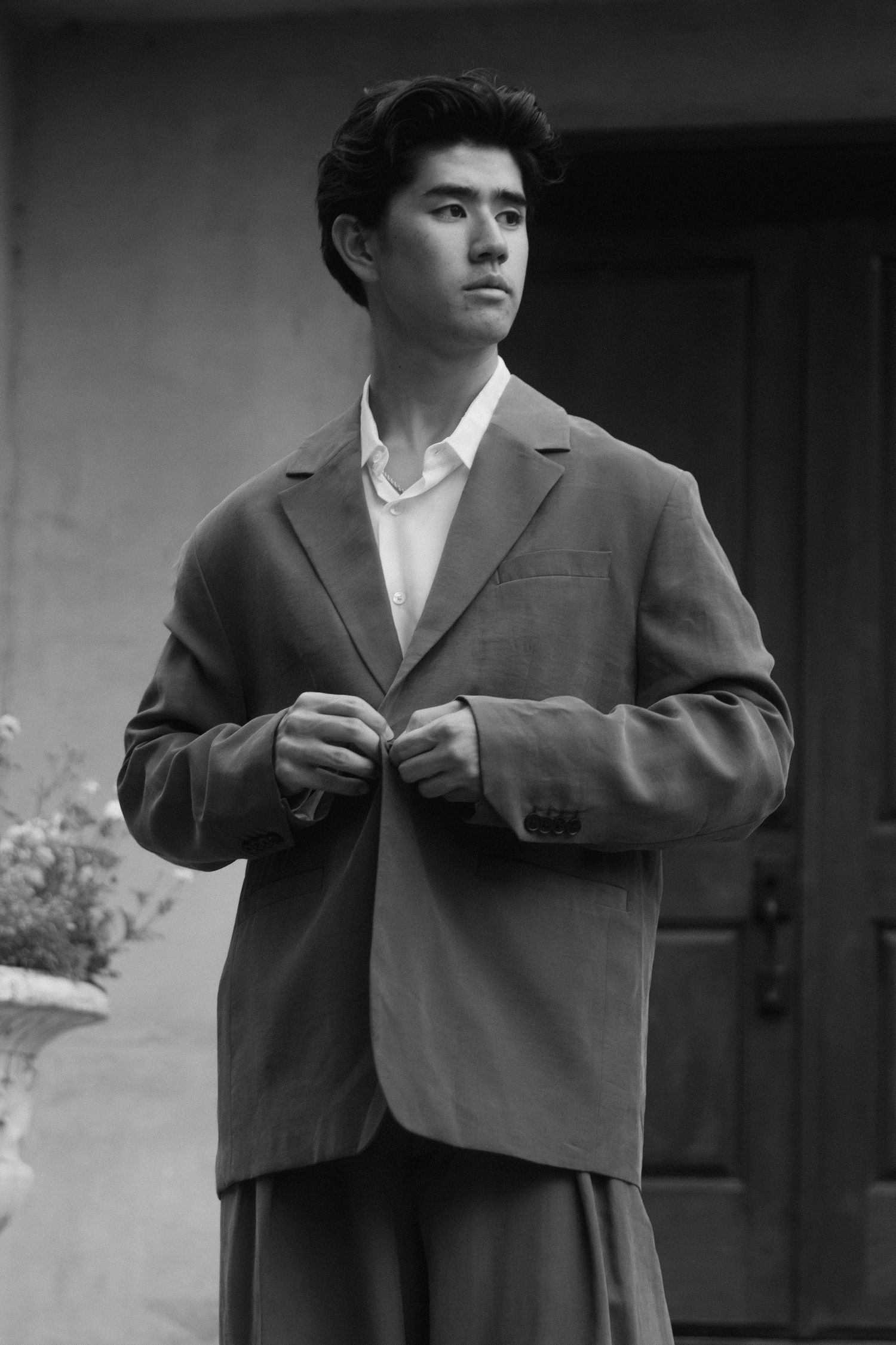 Young man in oversized blazer and white shirt buttoning jacket outdoors in black and white photo.