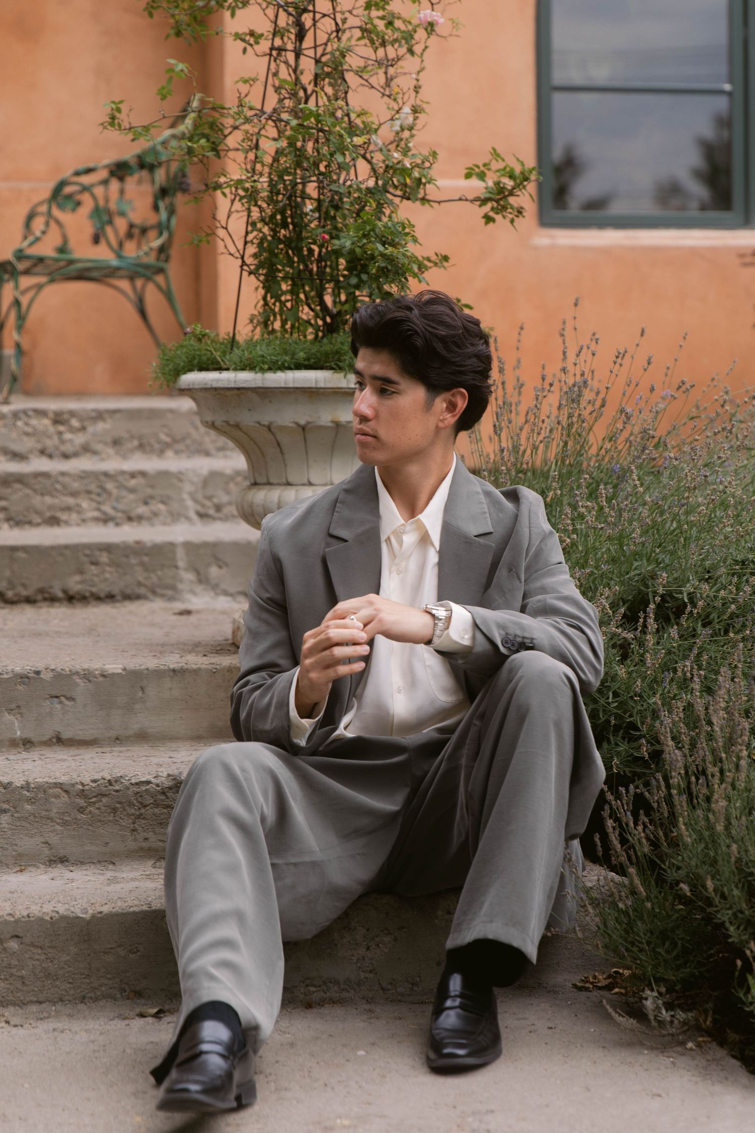 Young man in gray suit sitting on stone steps beside lavender plants near a terracotta building.