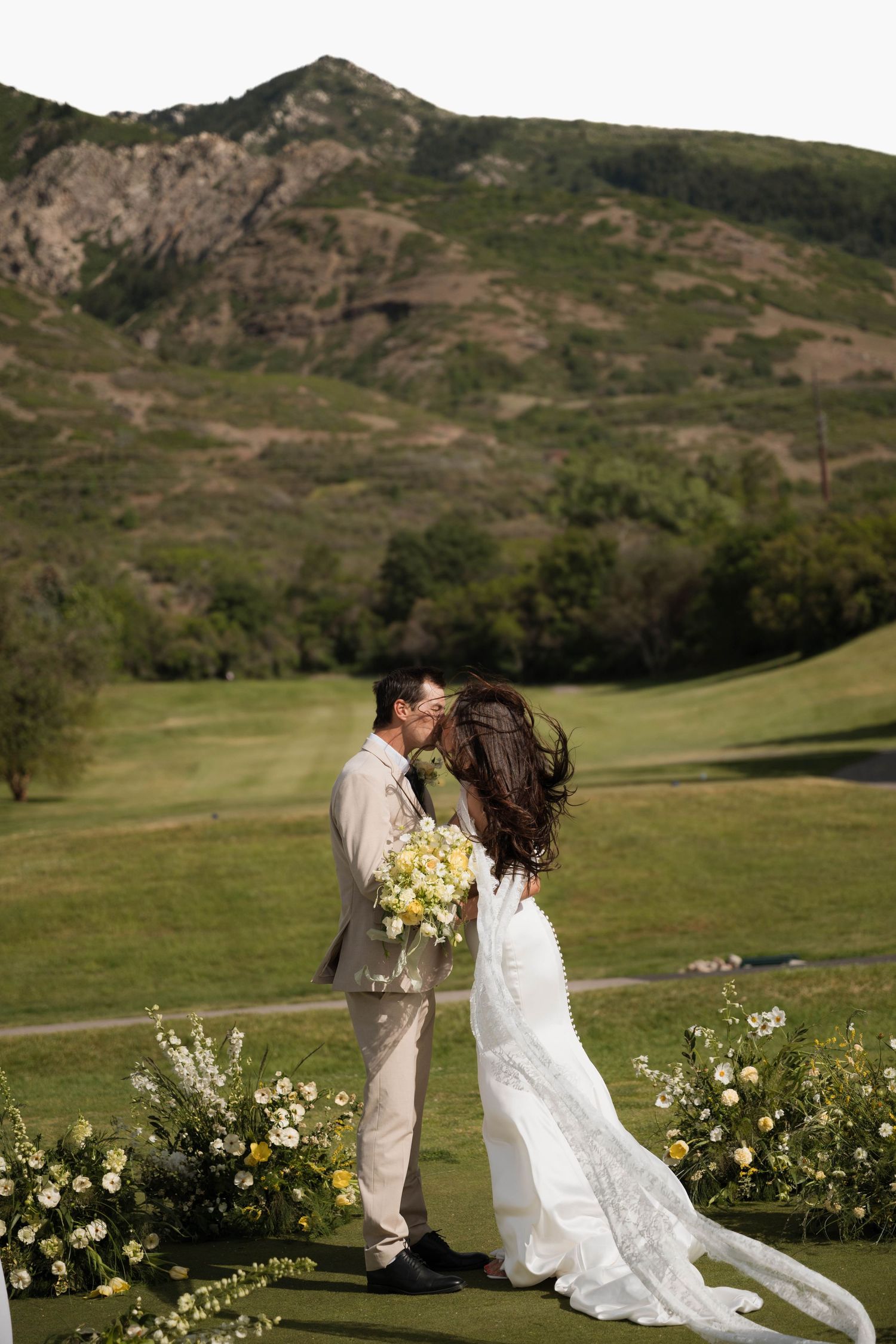 Bride and groom share first kiss at outdoor golf course ceremony with mountain views and floral decor.