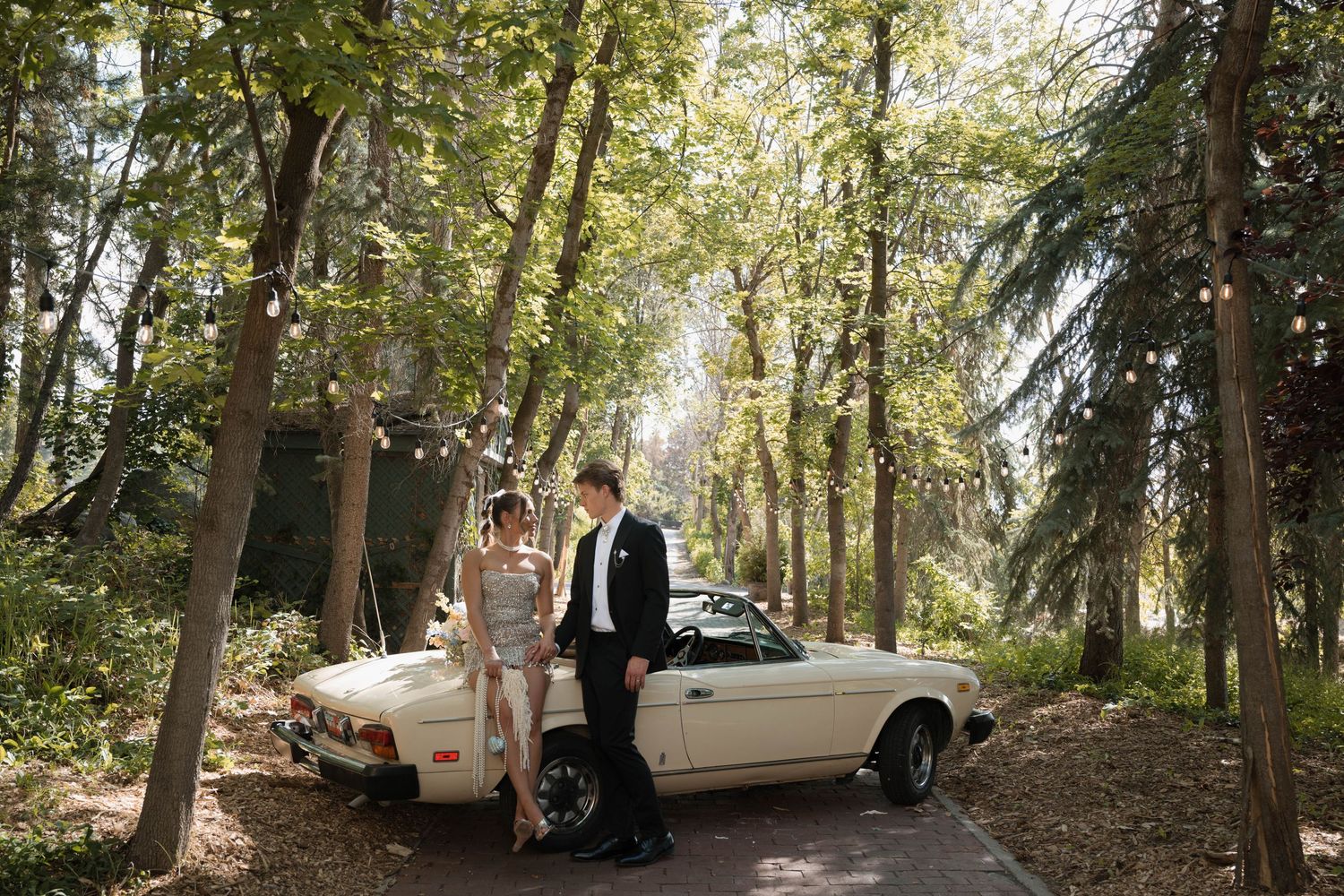 Bride and groom stand beside a vintage white convertible car on a tree-lined path with string lights.