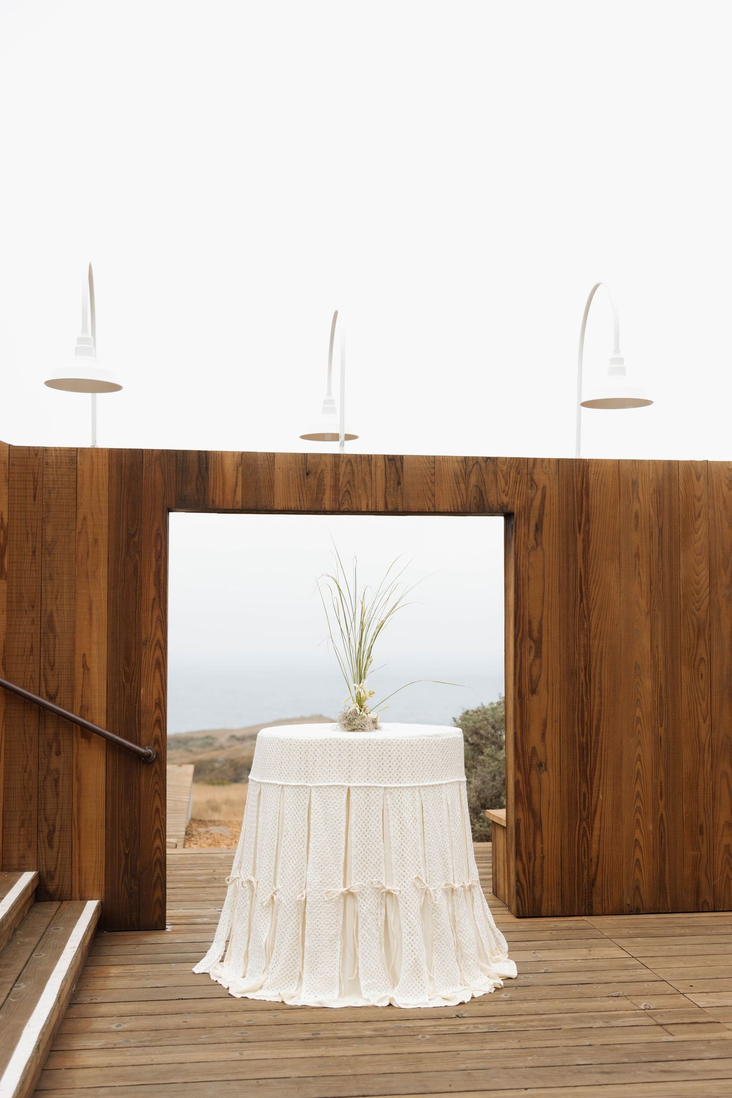 Elegant white draped table with plant centerpiece on wooden deck overlooking ocean, framed by modern wood walls.