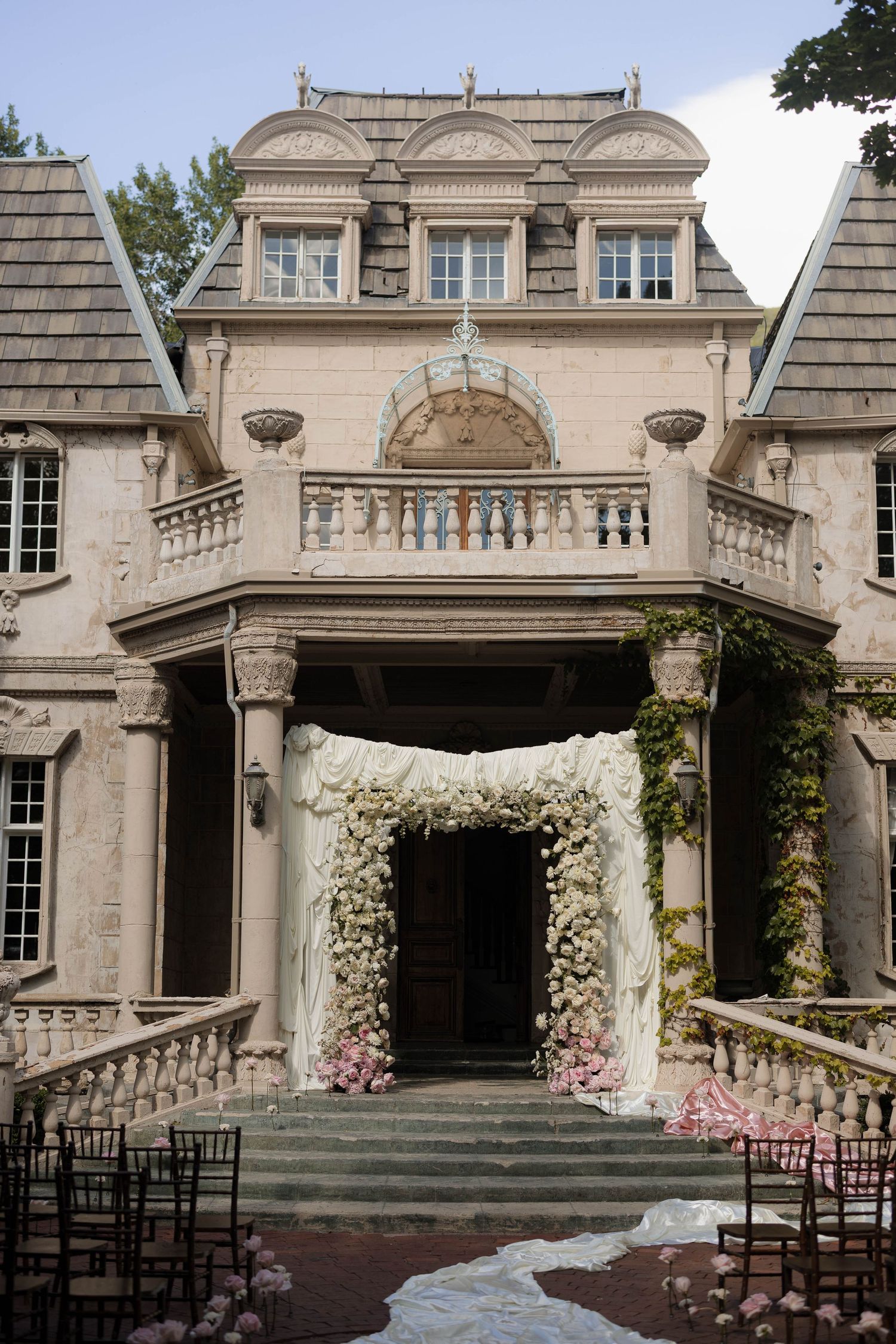 Elegant mansion entrance decorated with white floral arch and pink flowers for a wedding ceremony.